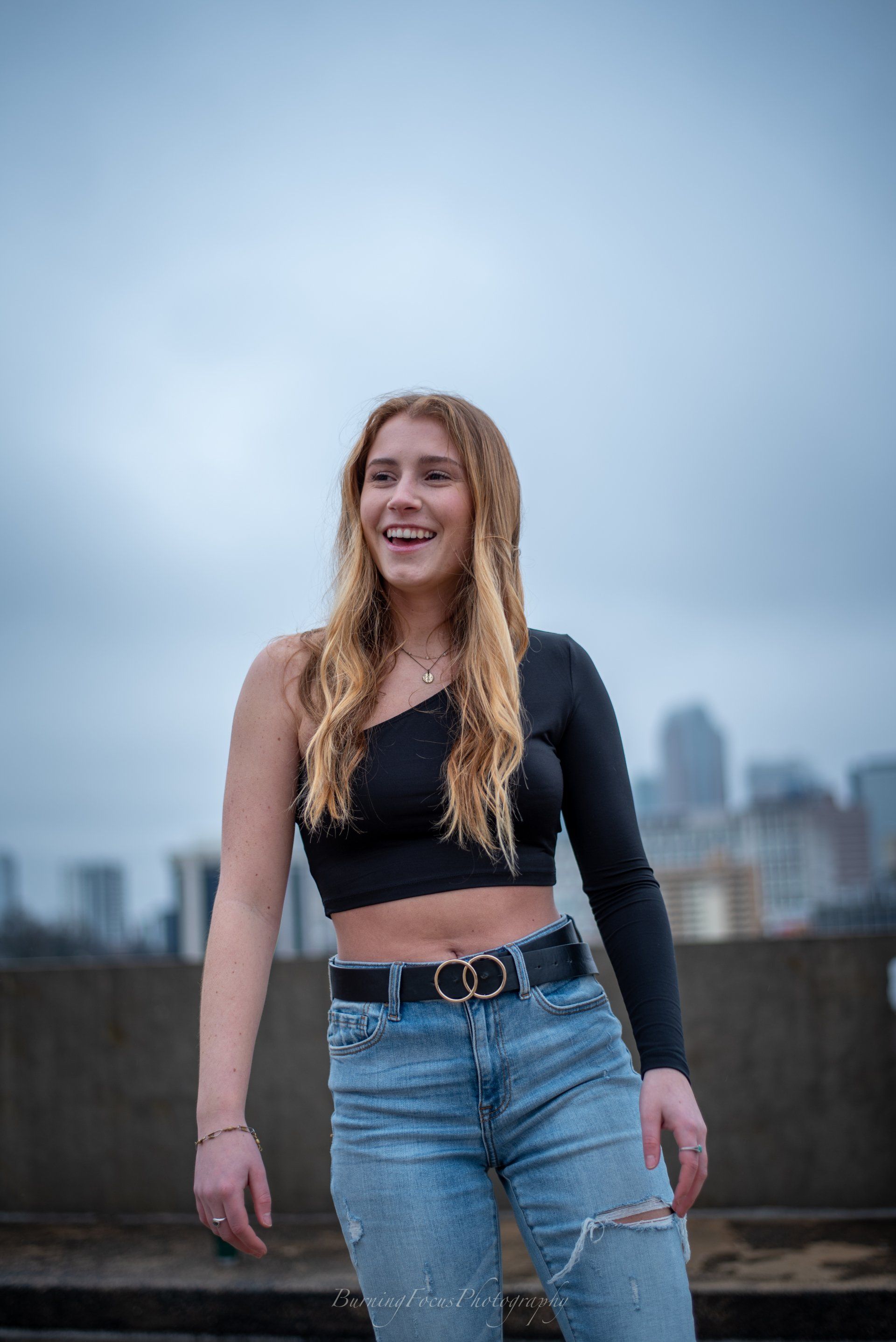 A woman in a black crop top and blue jeans is standing in front of a city skyline.