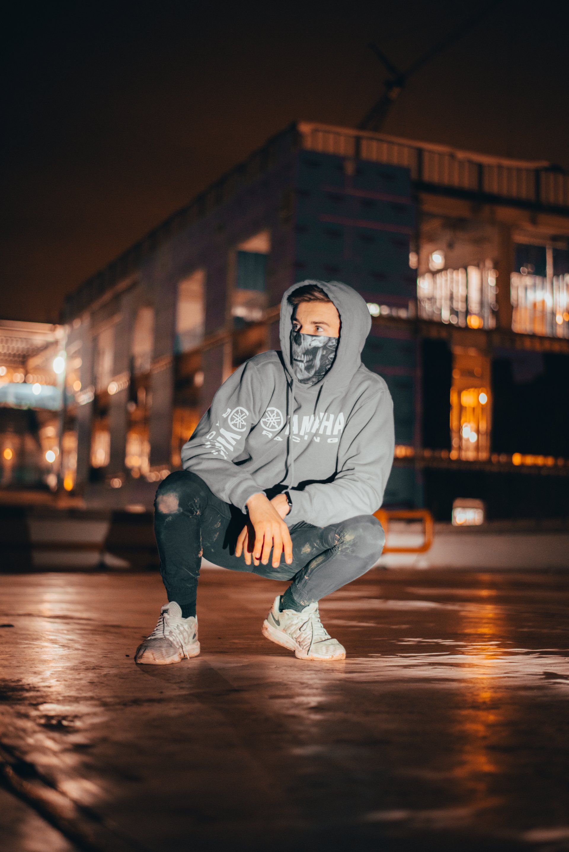 A man wearing a hoodie and a mask is squatting down in front of a building.