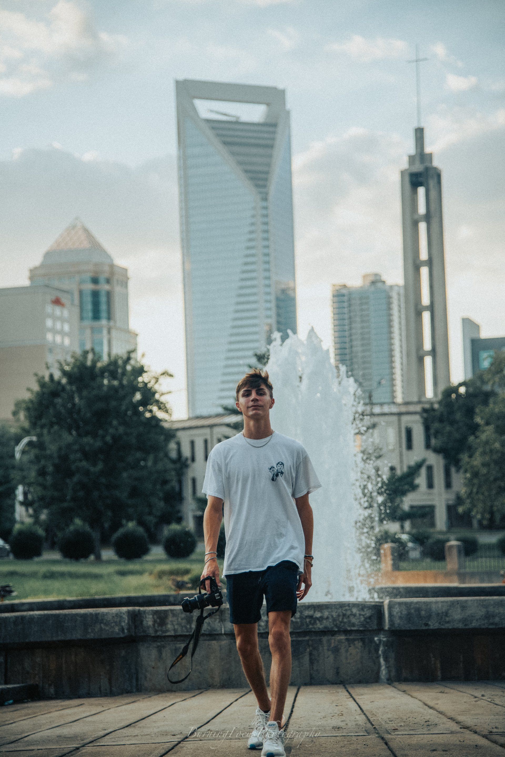A man is standing in front of a fountain in a city holding a camera.