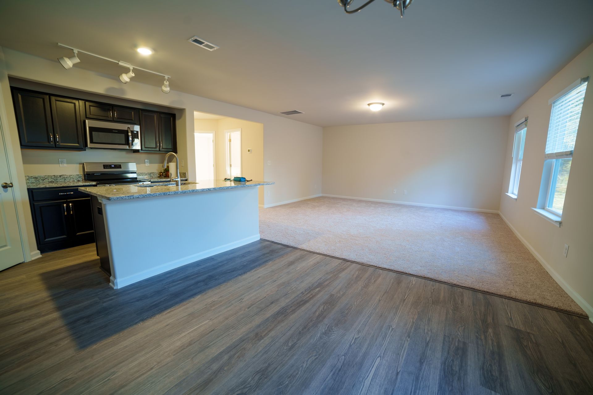 An empty living room with a kitchen and a large carpeted floor.