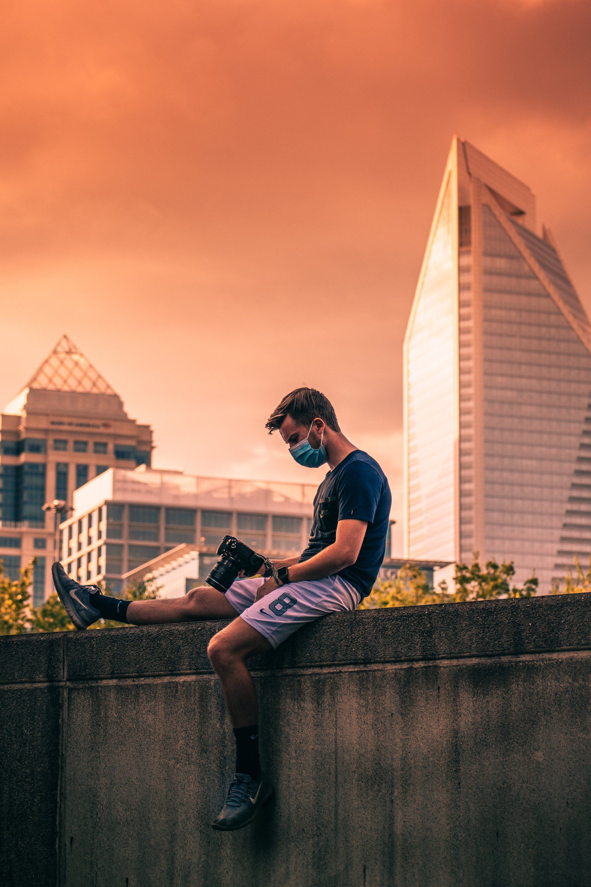 A man wearing a mask is sitting on a wall with a camera.