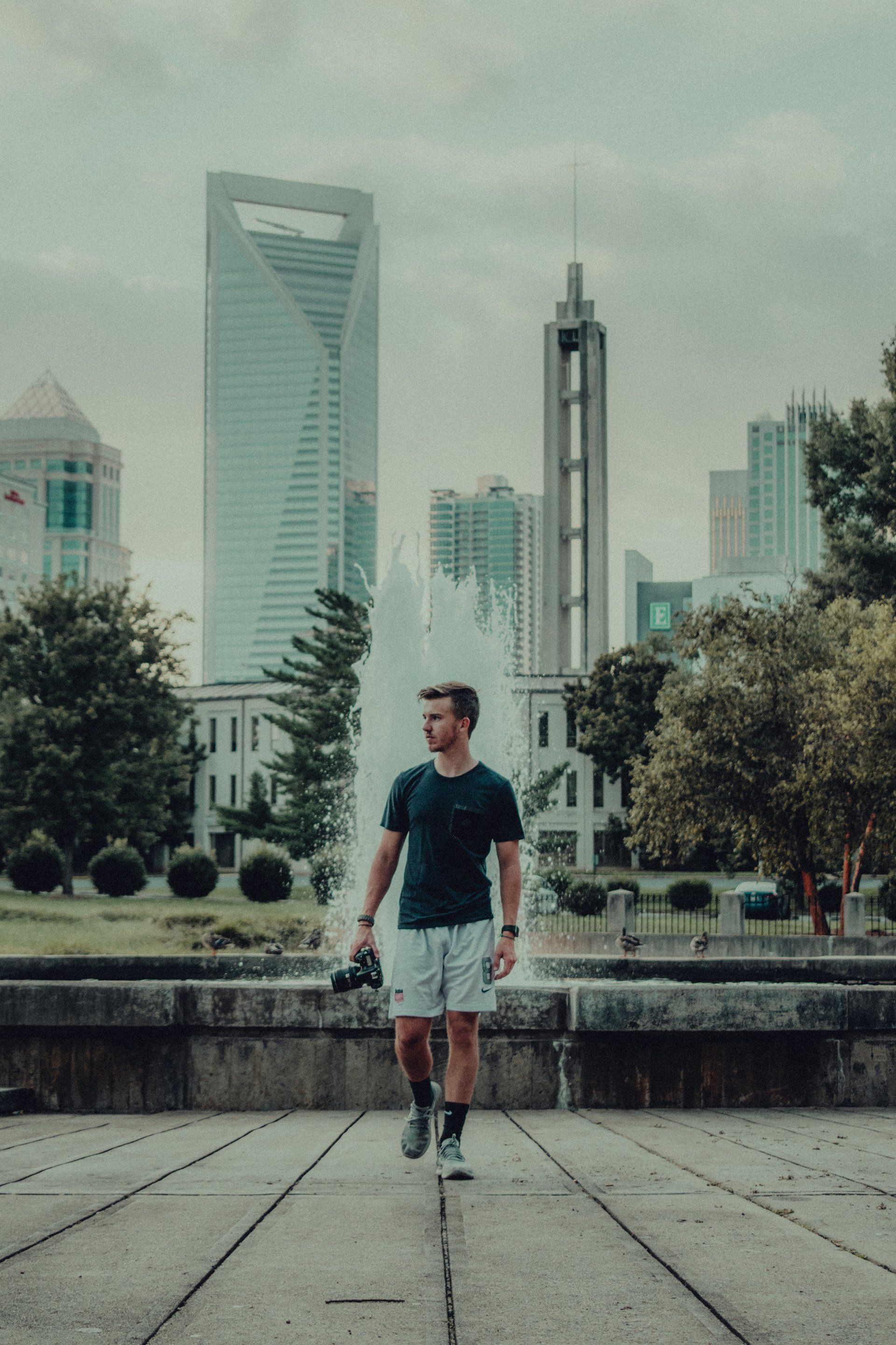 A man is walking in front of a fountain in a city park.