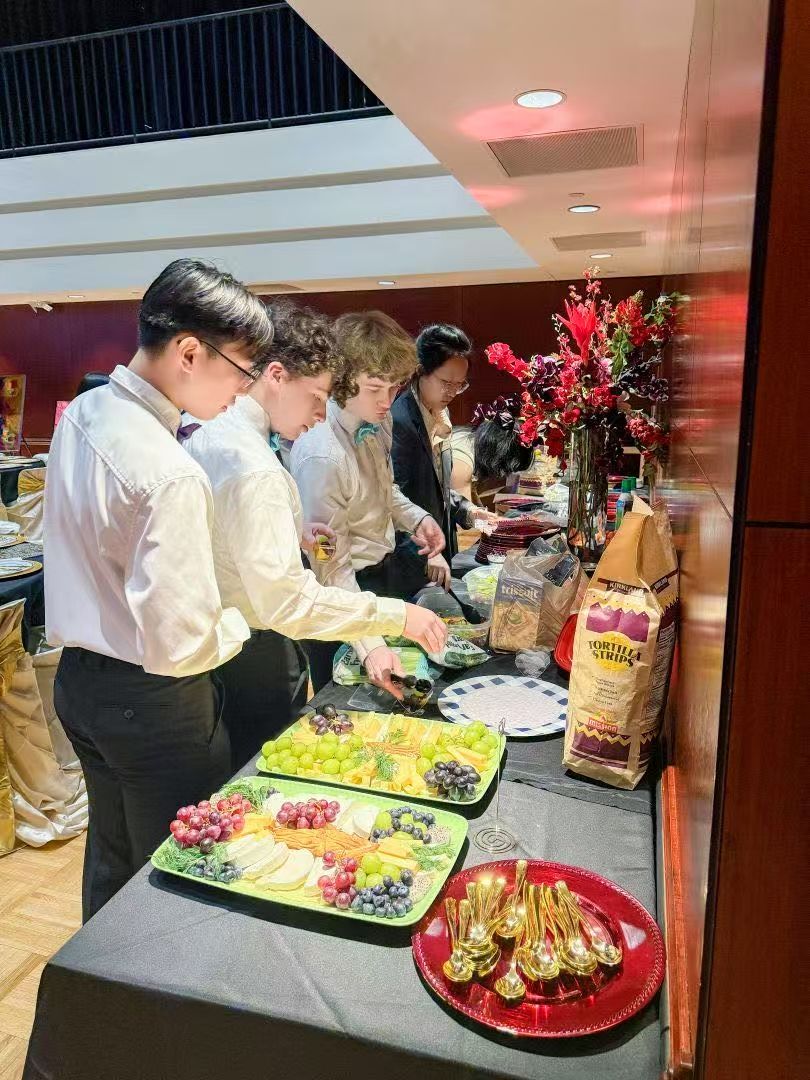 People at a buffet table, selecting food. Fruit platters and a floral arrangement visible.