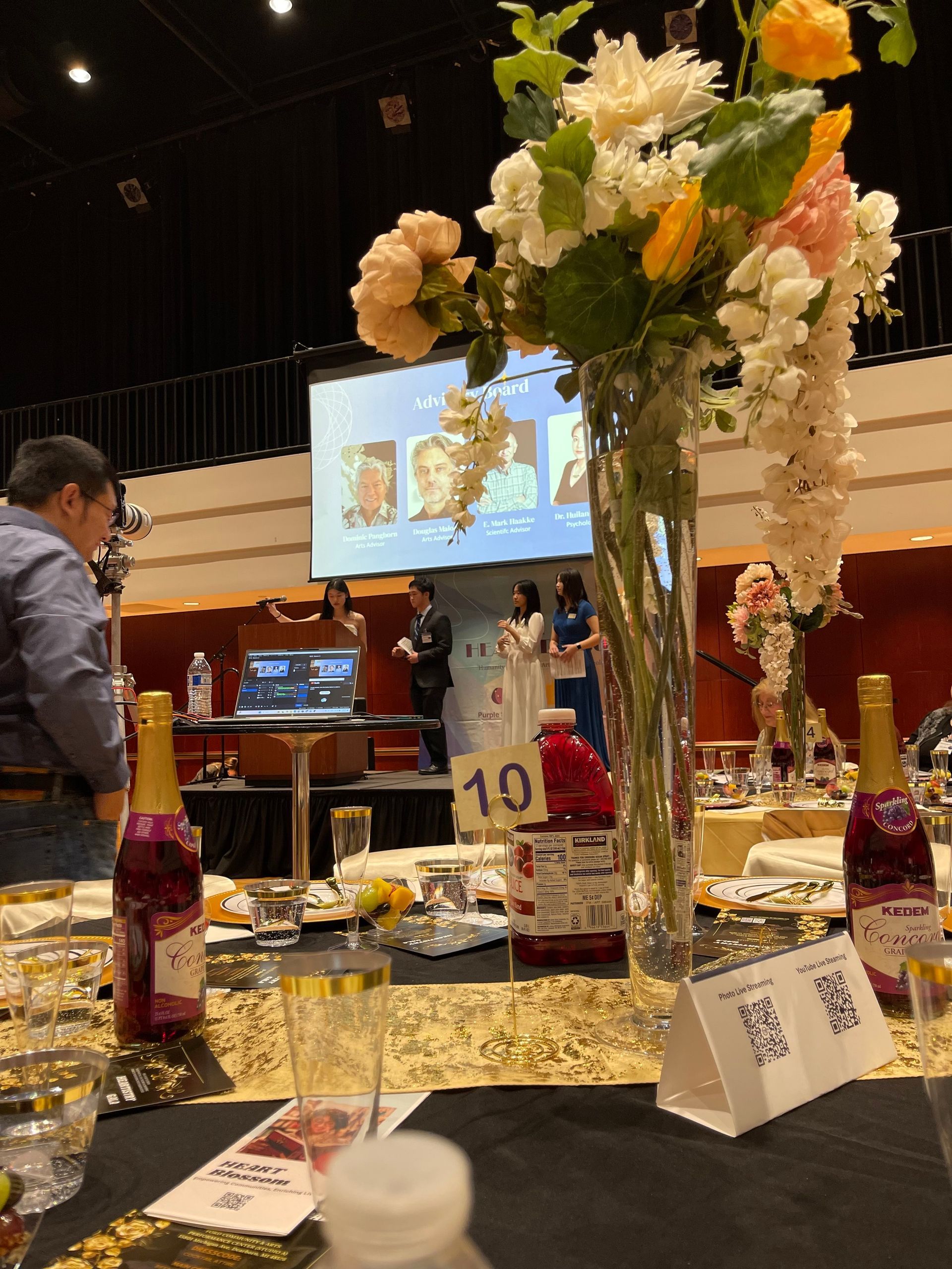 Formal event with speakers on stage, decorated tables, floral centerpiece, and wine bottles.