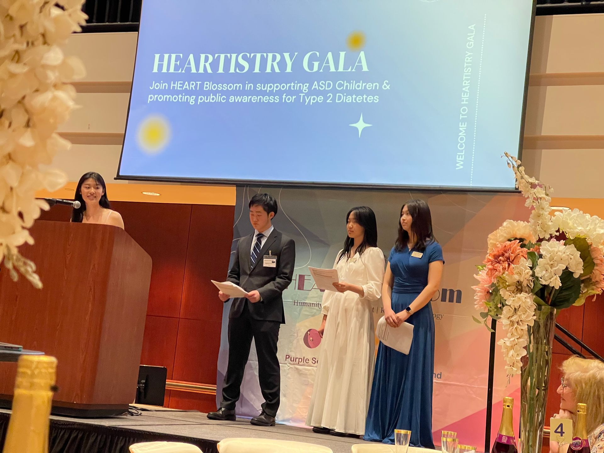 Heartistry Gala event: speakers on stage, woman at podium, others holding papers, banner.