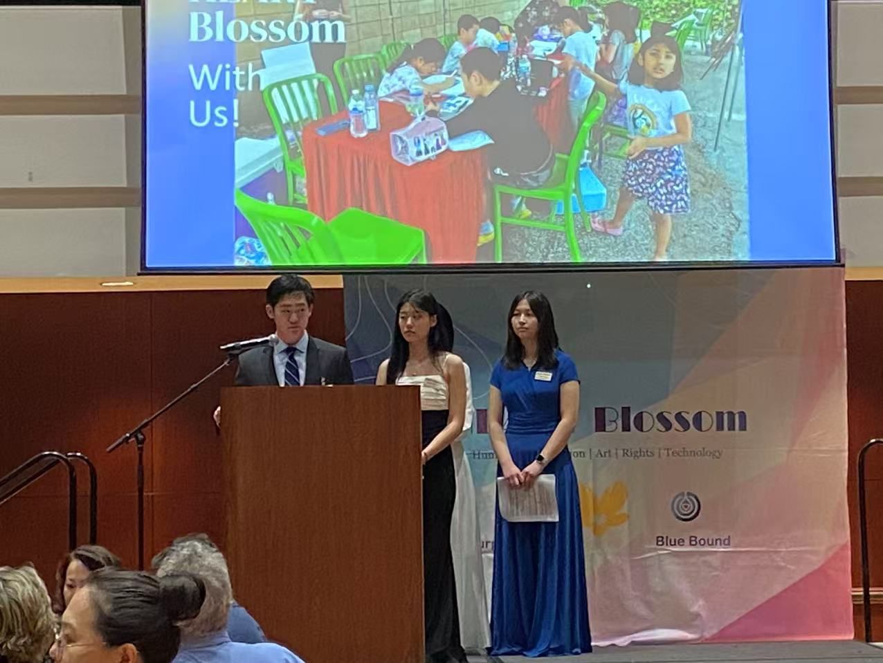 Three people presenting at a Blossom fundraising event. A screen shows children at tables; audience visible.