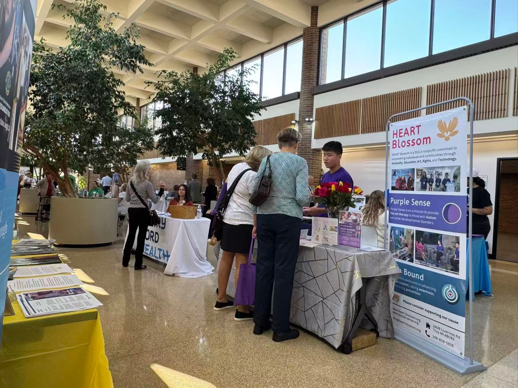 People at a booth, likely an event. Indoor setting with a banner, trees, and information on tables.