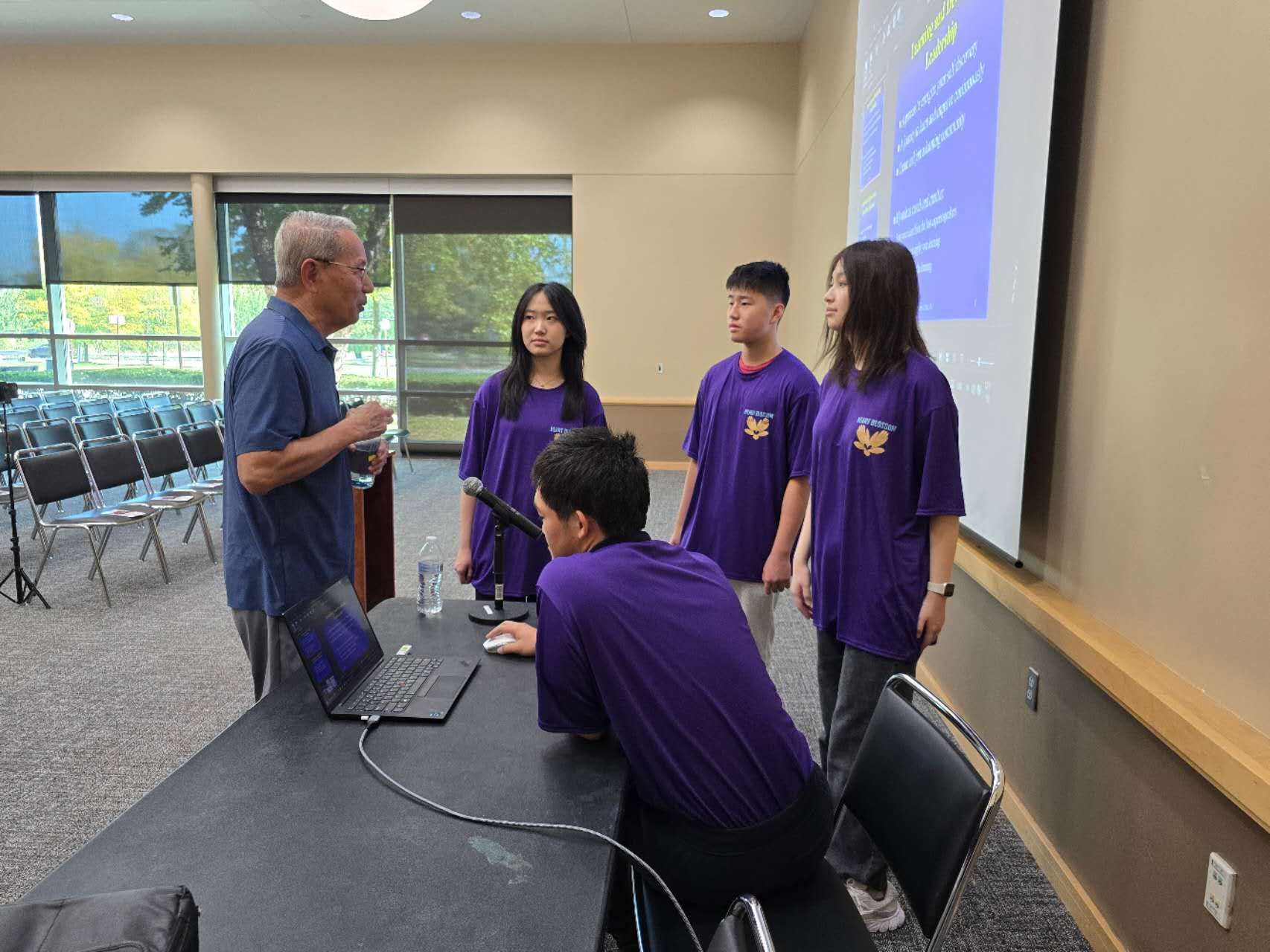 A man speaks to four people wearing purple shirts at a table with a laptop and a projector screen.