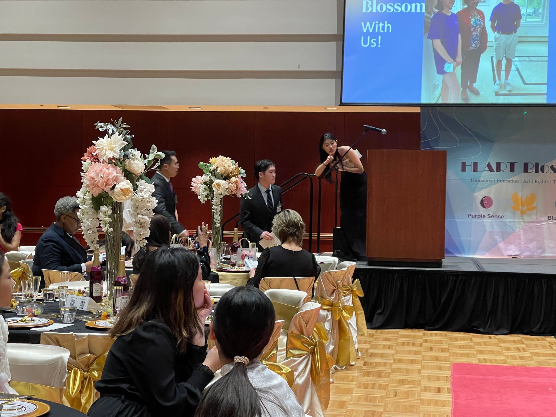 Woman speaking at a podium on stage at a gala. Guests seated at tables with floral pieces. 