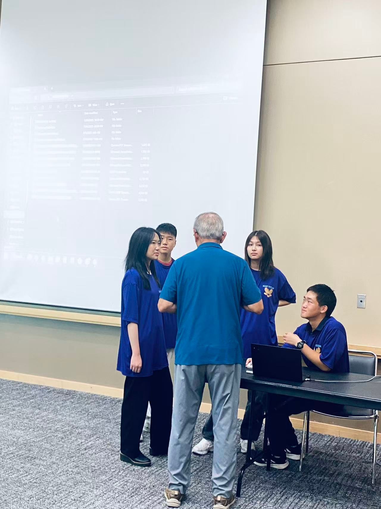 A group of people in blue shirts and pants talking in a room, a man standing in front of them.