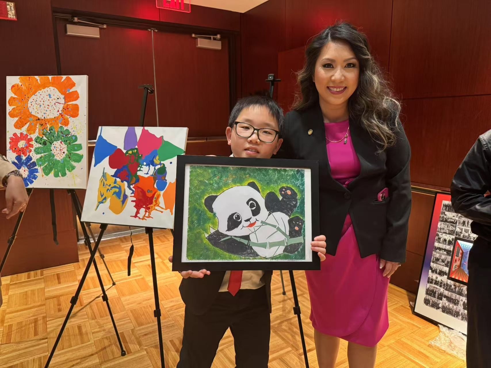 A young boy holding a framed panda painting stands with a woman, both smiling, next to artwork on easels in a room.