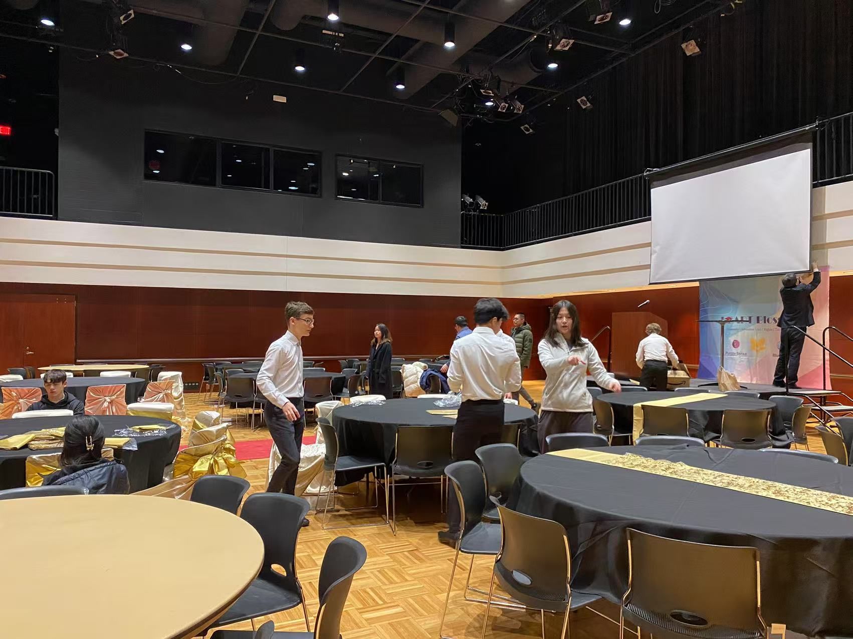 People setting up tables and equipment in a large event hall.
