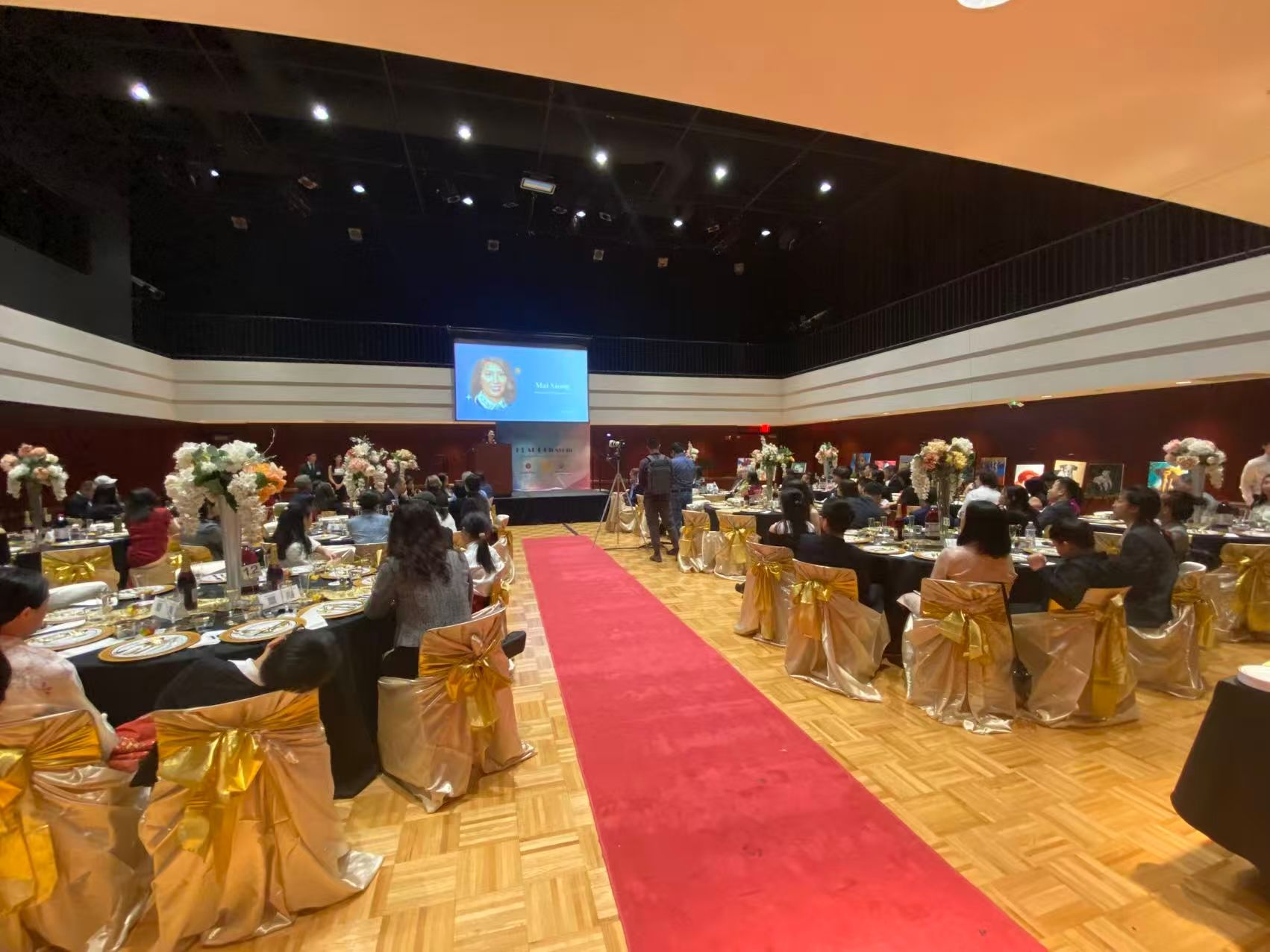 An event in a large ballroom with tables set, red carpet, people seated, and a presentation screen.