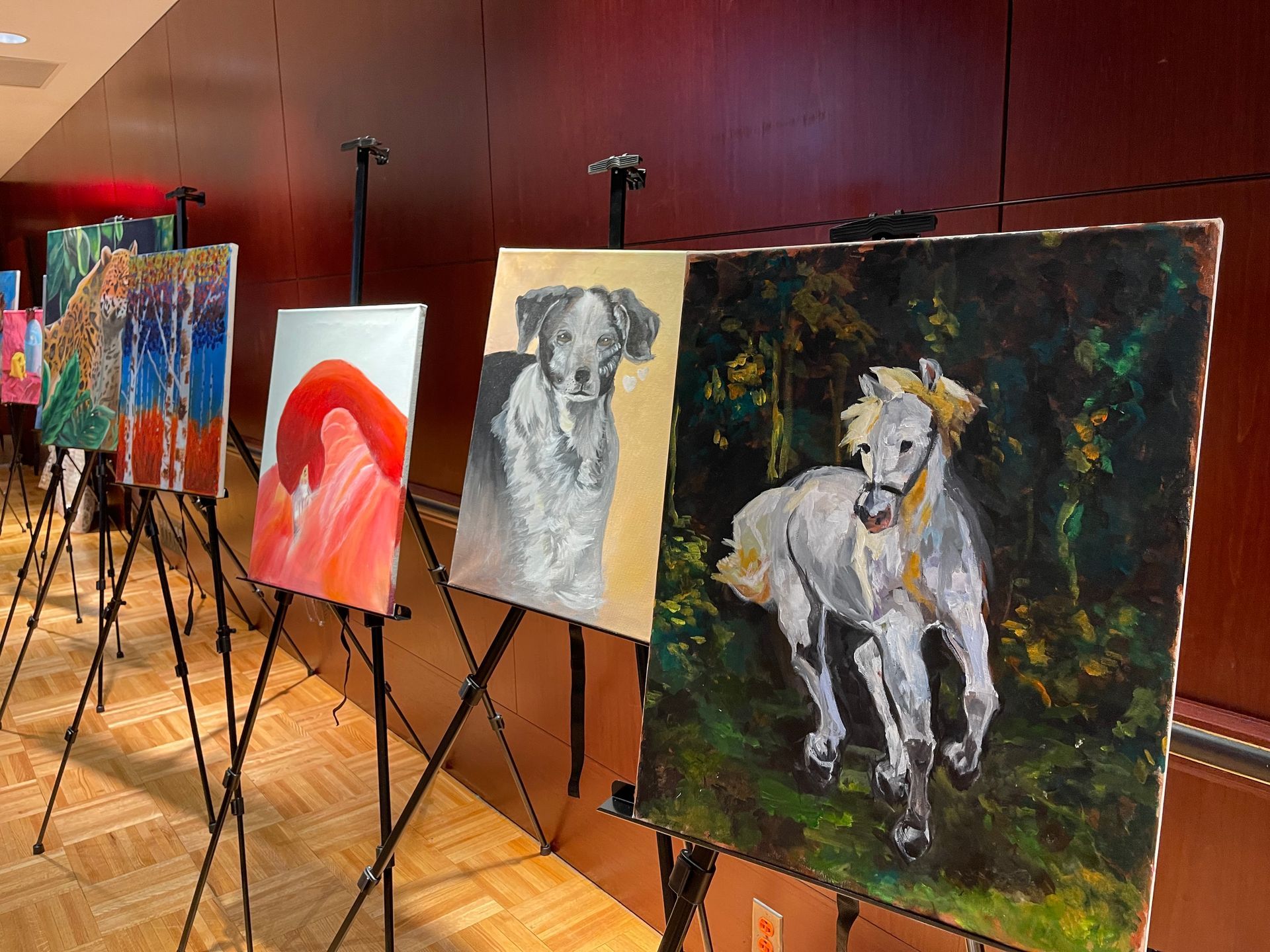Paintings on easels in a row, showcasing animal portraits and landscapes, displayed against a dark wall.