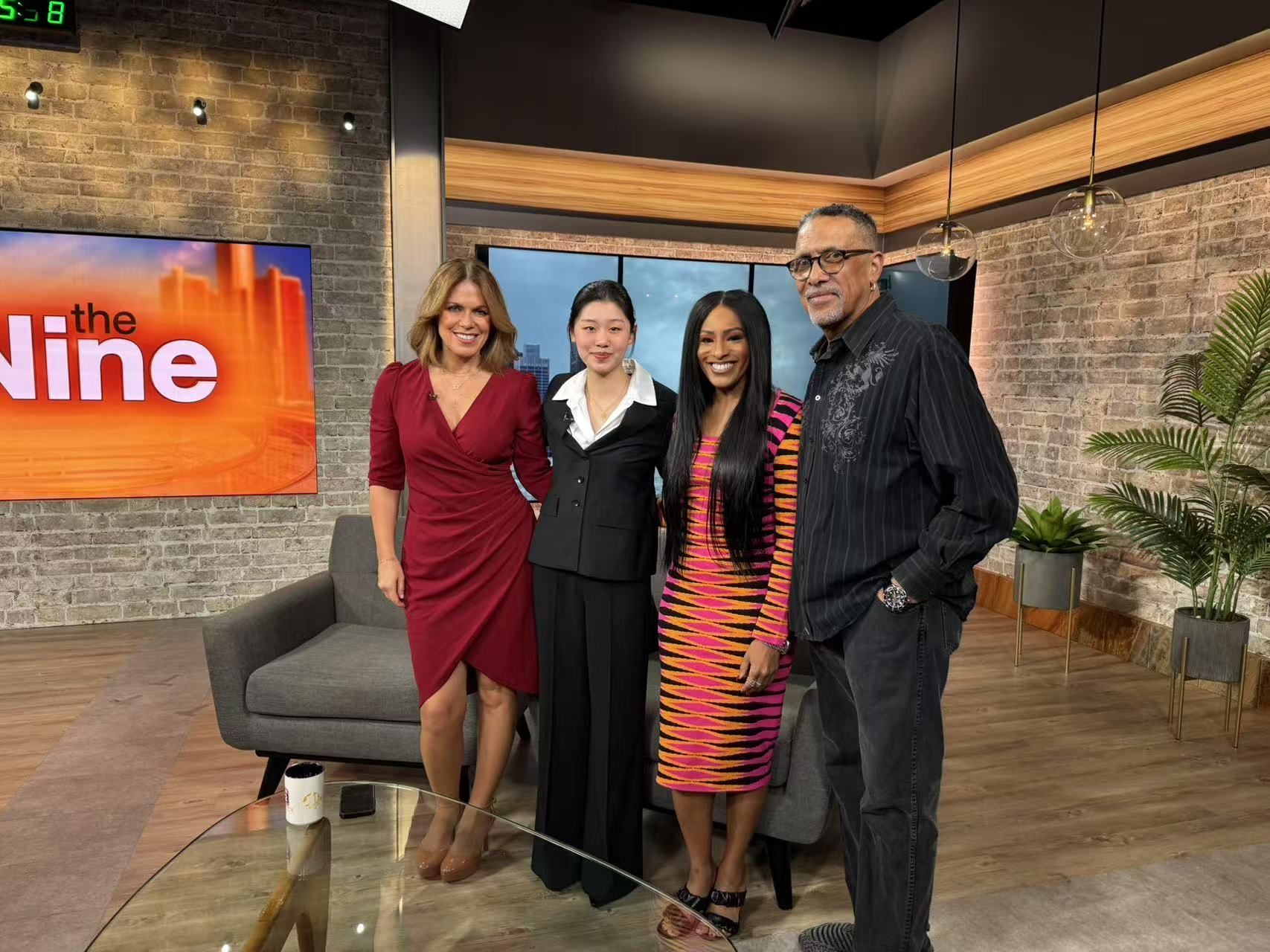 Four people pose for a photo in a TV studio. The set includes a couch and plants.