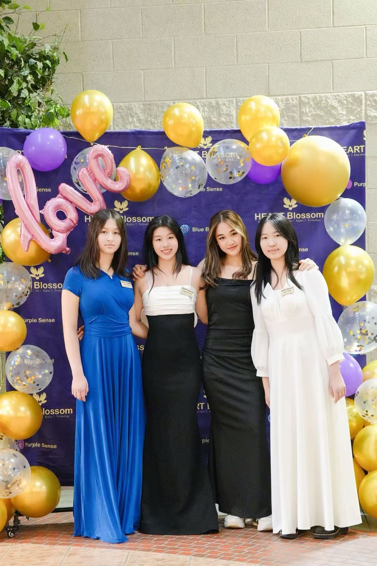 Four young women in formal dresses pose in front of a decorated backdrop with balloons.