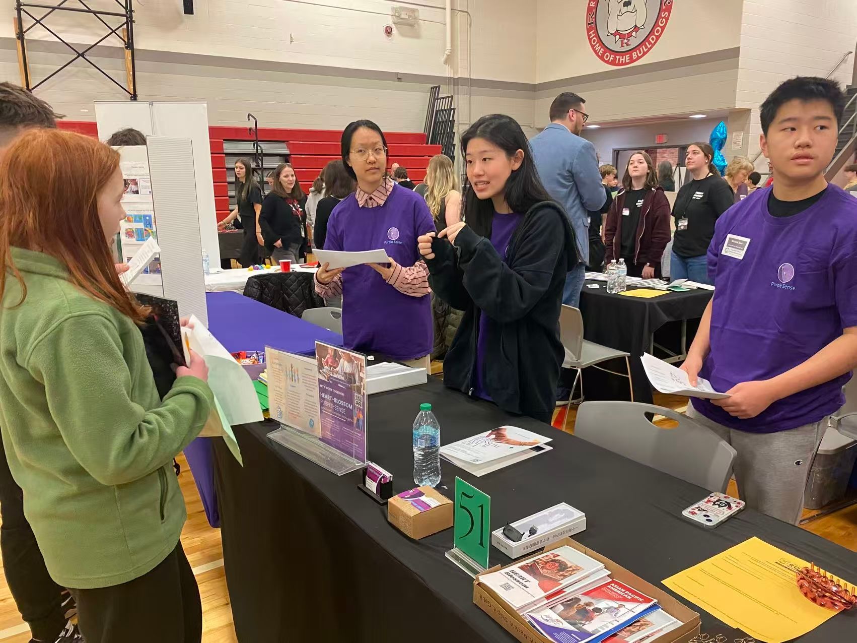 Students at a table in a gymnasium, talking. Several wear purple shirts. Flyers