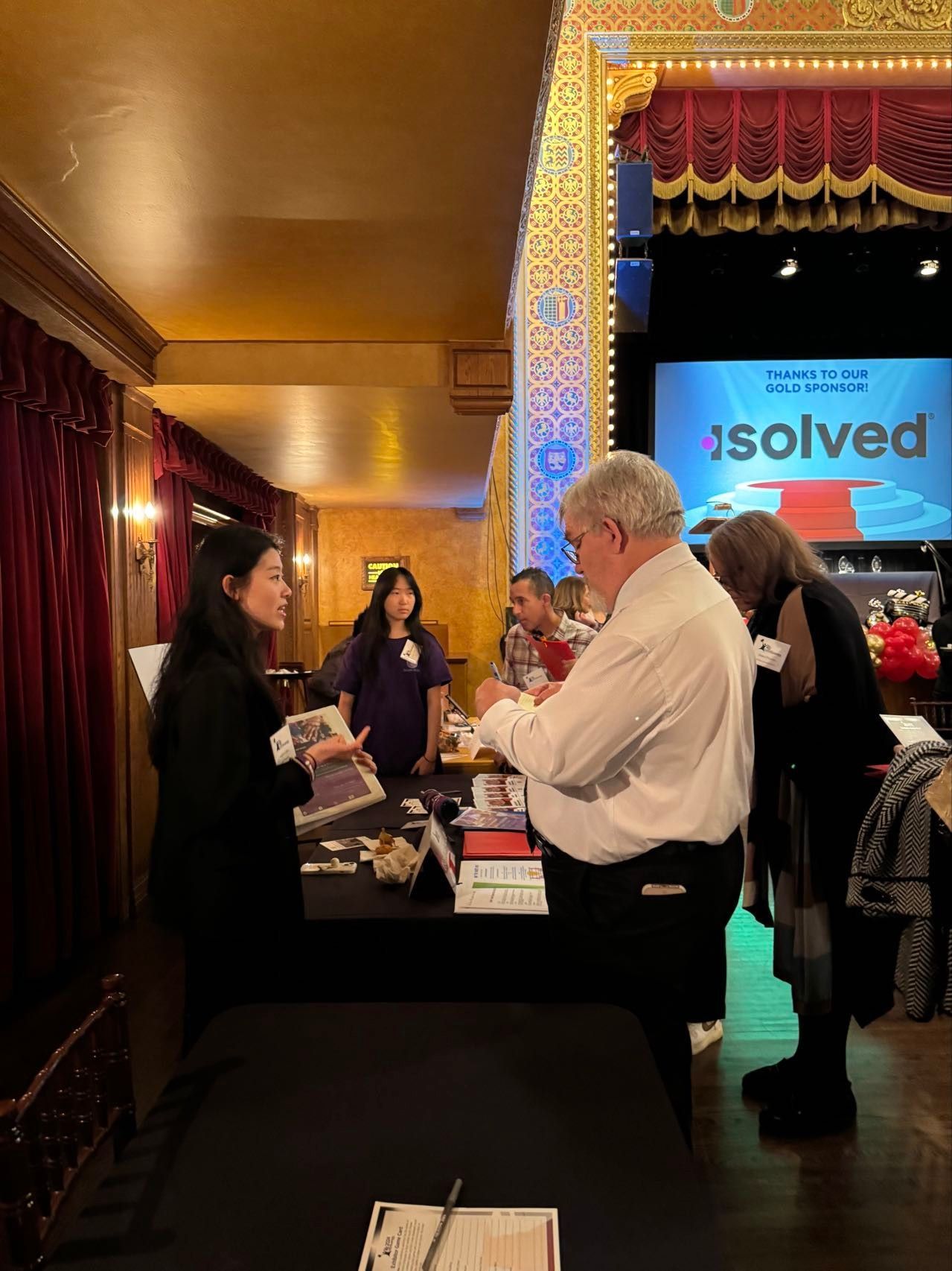 People at a booth, attending an event. Two women, two men talking at a table. Stage in the background.