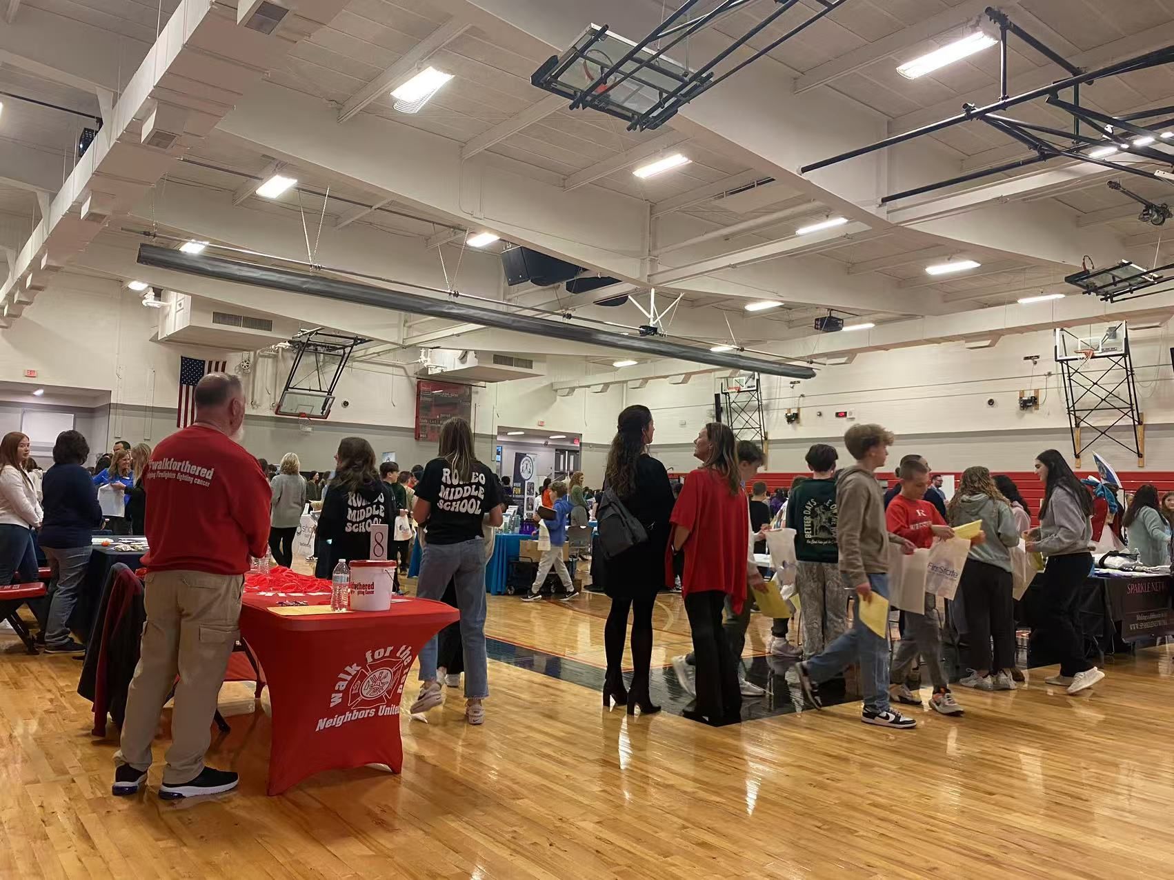 People gathered in a gymnasium, attending an event at tables. Red and white branding visible.