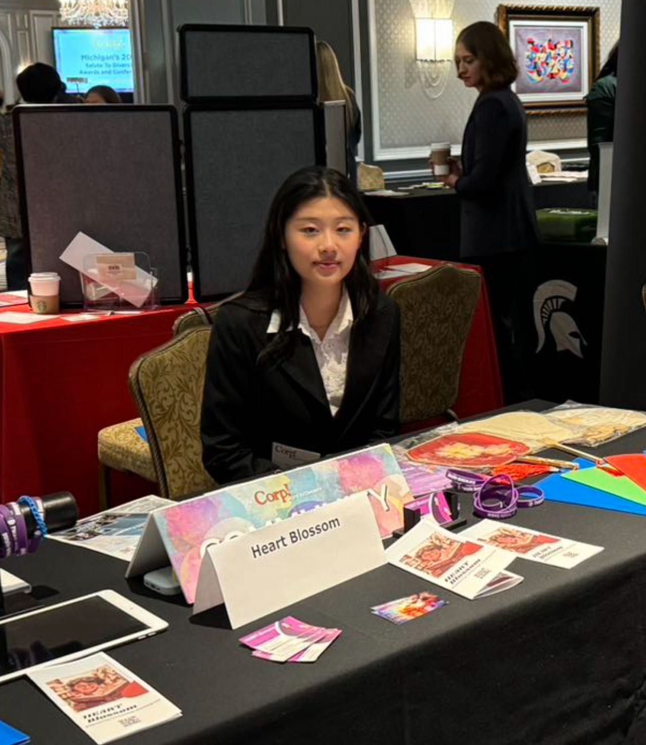 Young woman at a booth; wearing a black blazer; seated. Displays include business cards and colorful items, at a conference.
