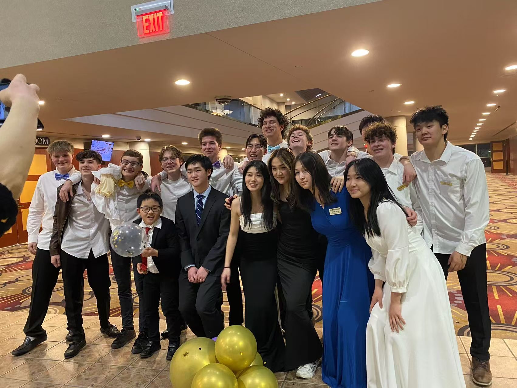 Group photo of teenagers in formal wear, smiling. Balloons on the floor, indoors, with an 