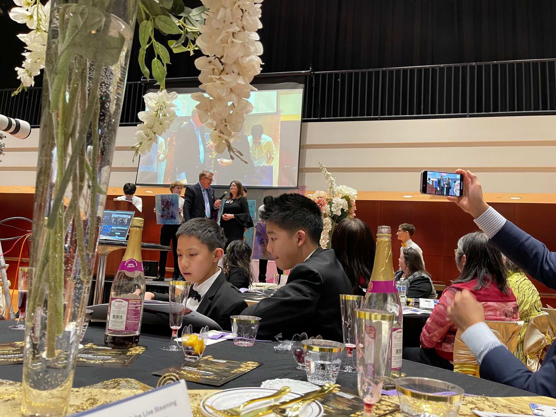 Formal event with people seated at tables, speakers on a stage, and someone recording with a phone.