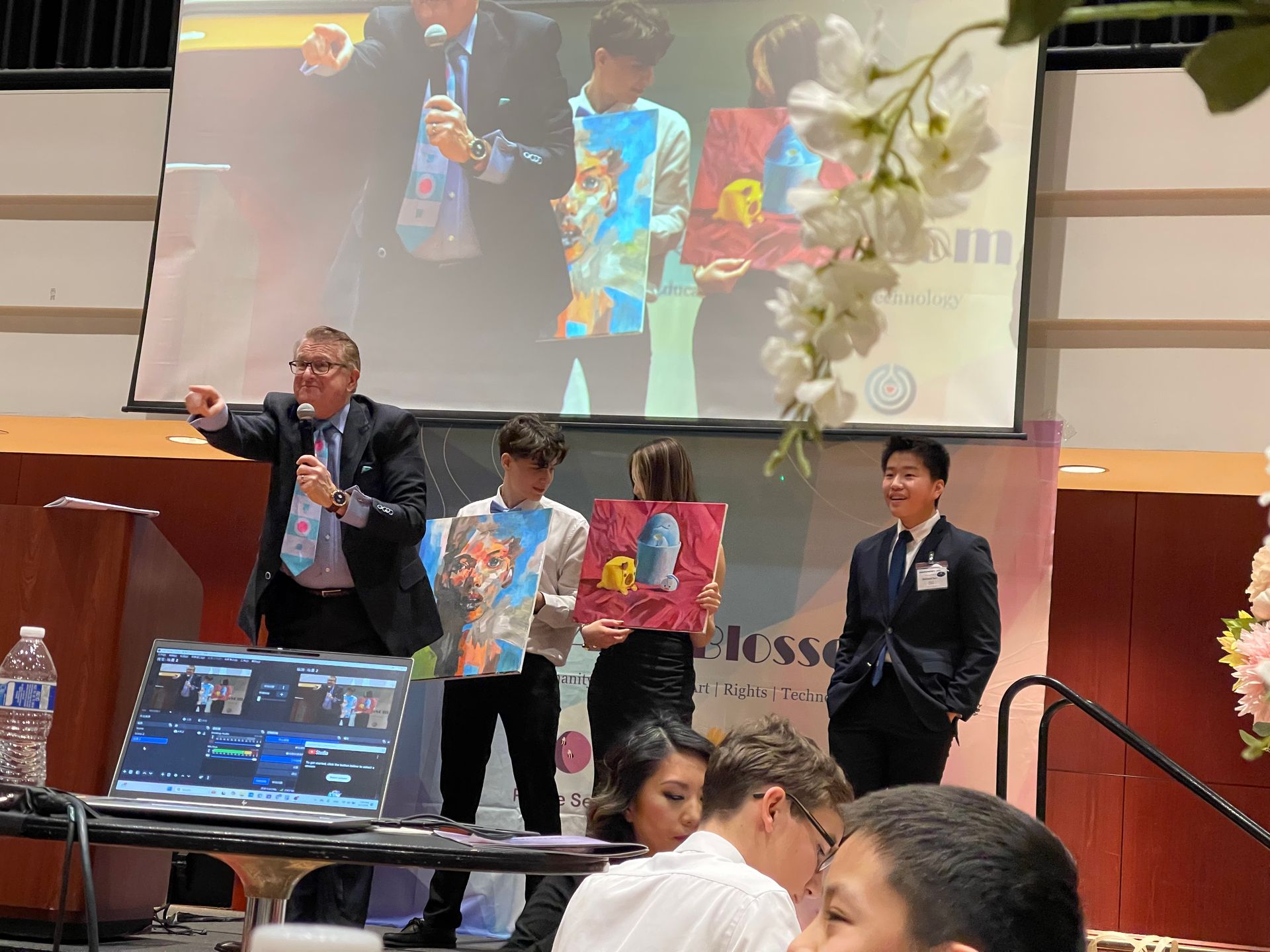 Man auctioning artwork on stage. Three people hold up art in front of a large screen. Audience seated below.