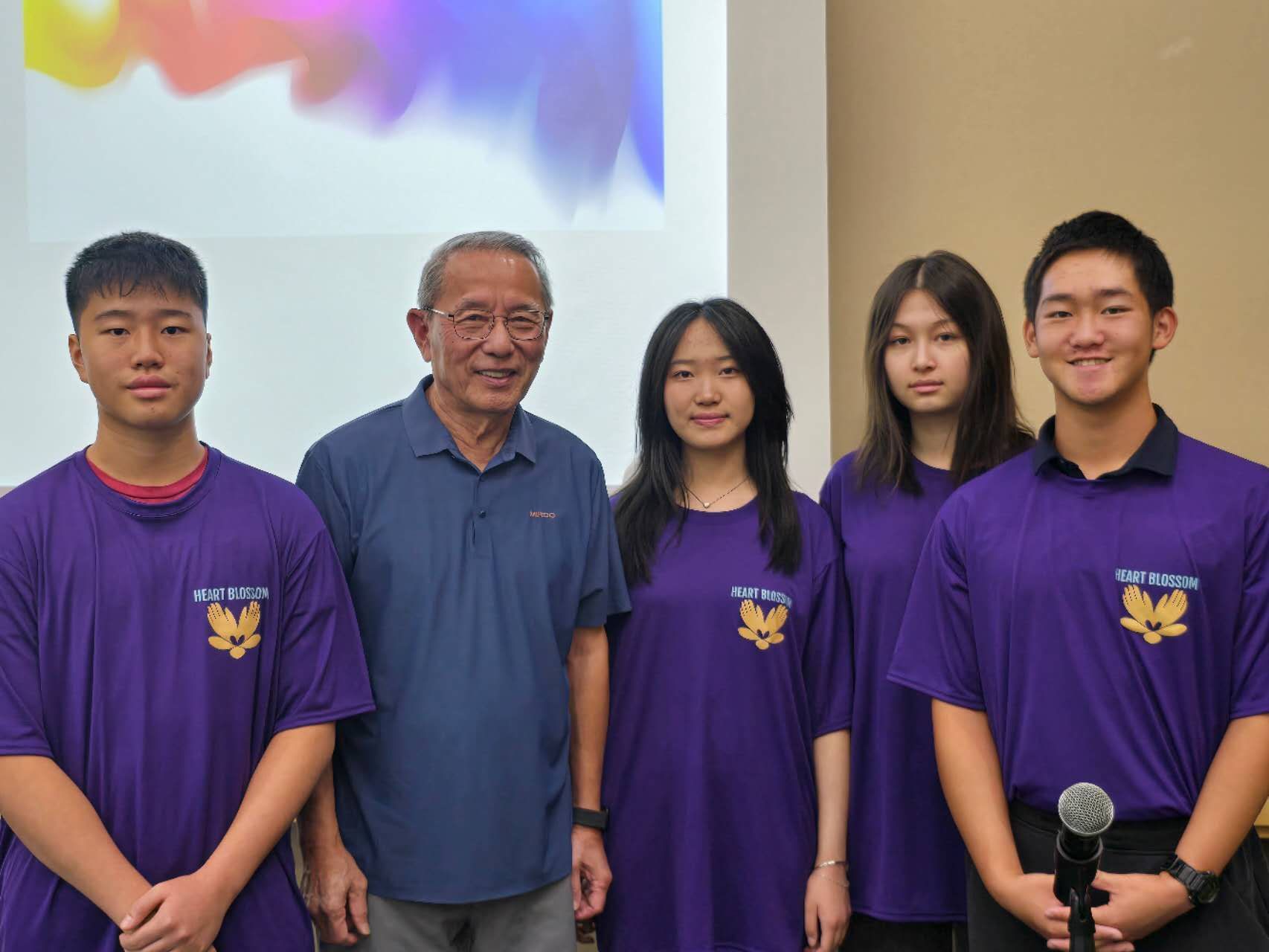 Five people pose. Man in blue shirt stands with four students in purple shirts. White background, colorful art.