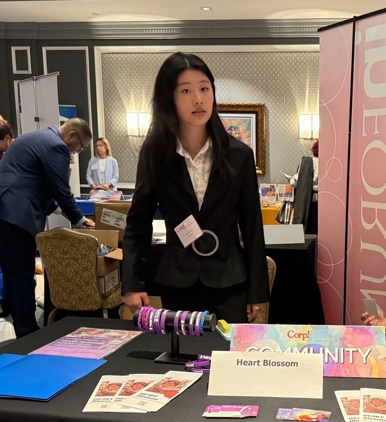 Woman in suit at a booth, holding a bracelet. Conference setting.