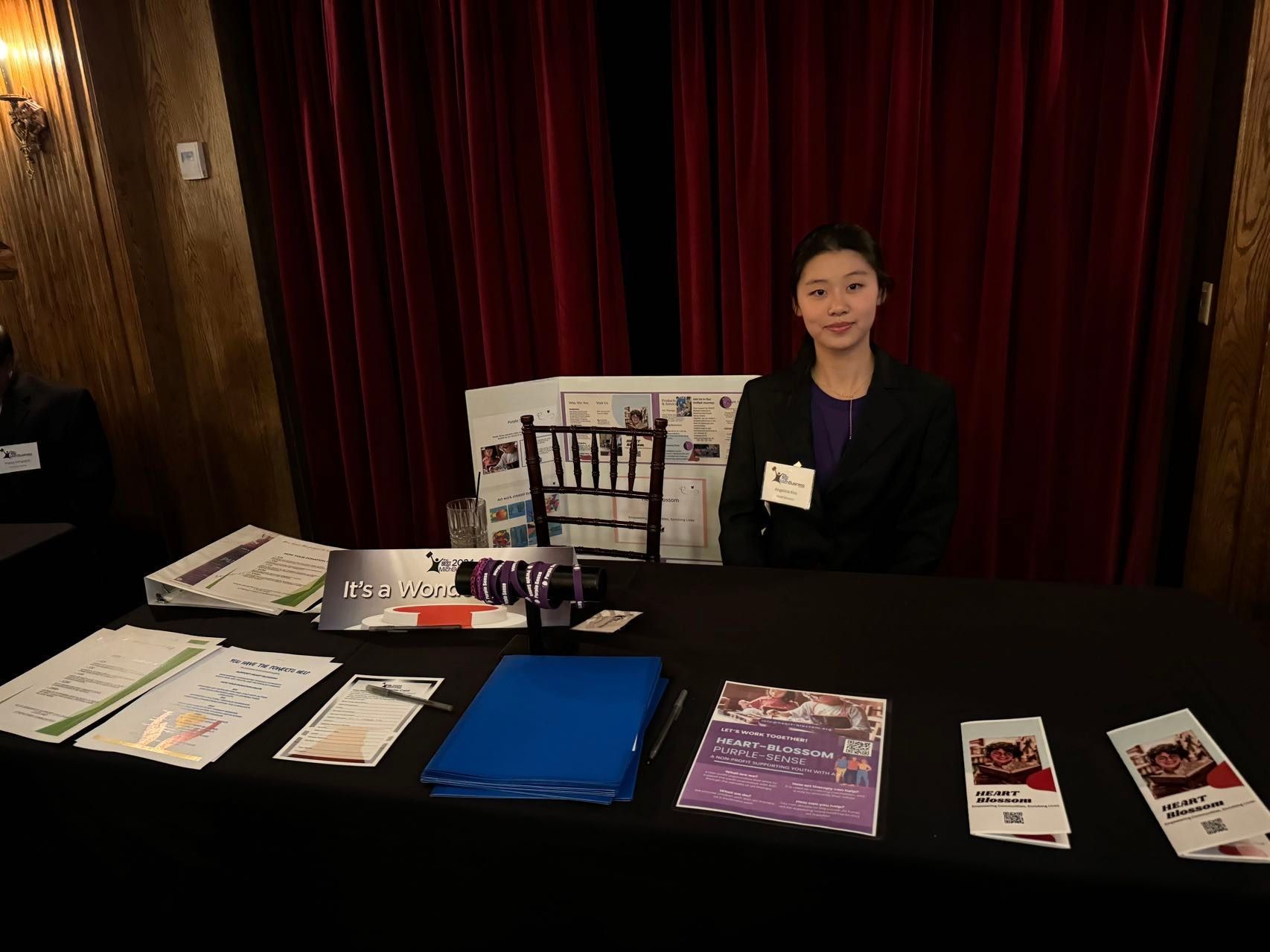 Woman at a table with brochures. Red curtain backdrop. She is wearing a black suit and smiling.