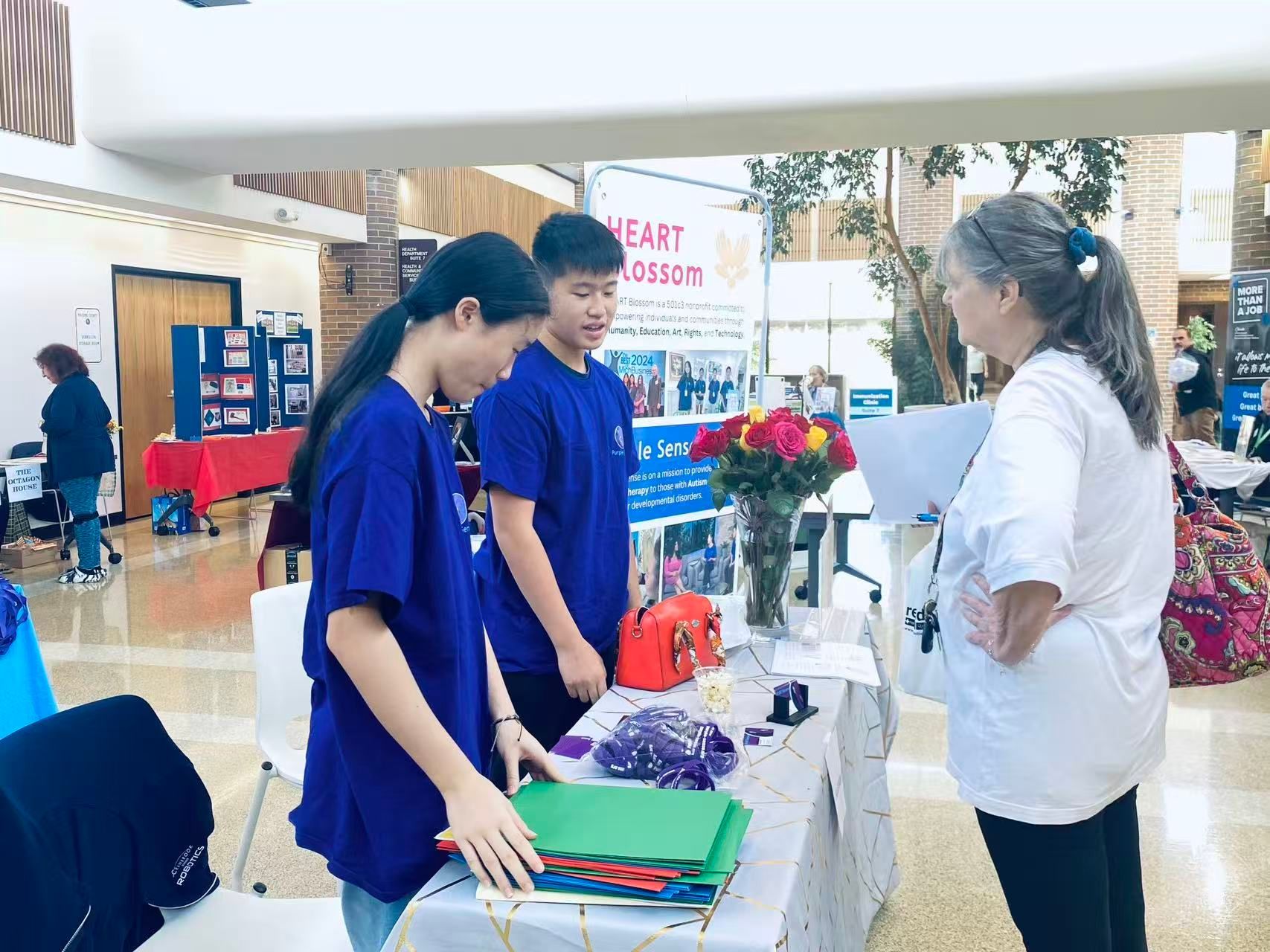 Two students in blue shirts at a table converse with an older woman, flowers in vase.