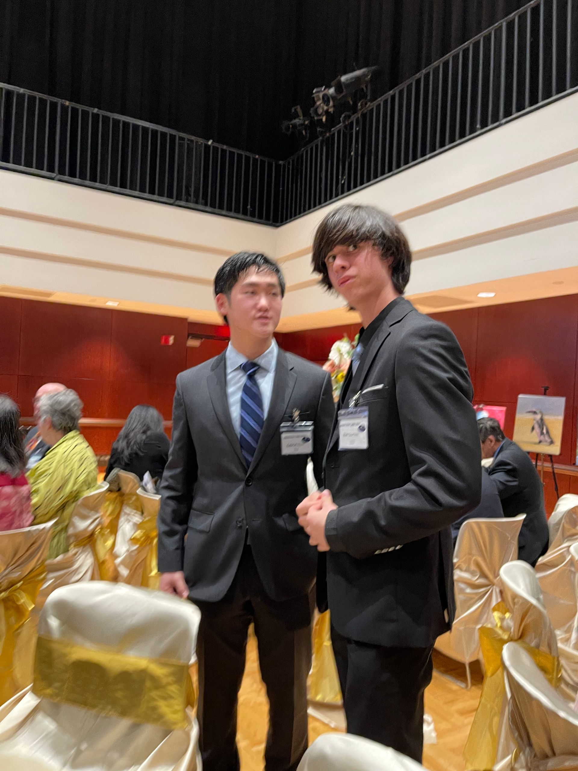 Two men in suits at a formal event. One is Asian, the other has long hair. Tables with gold covers are in the background.