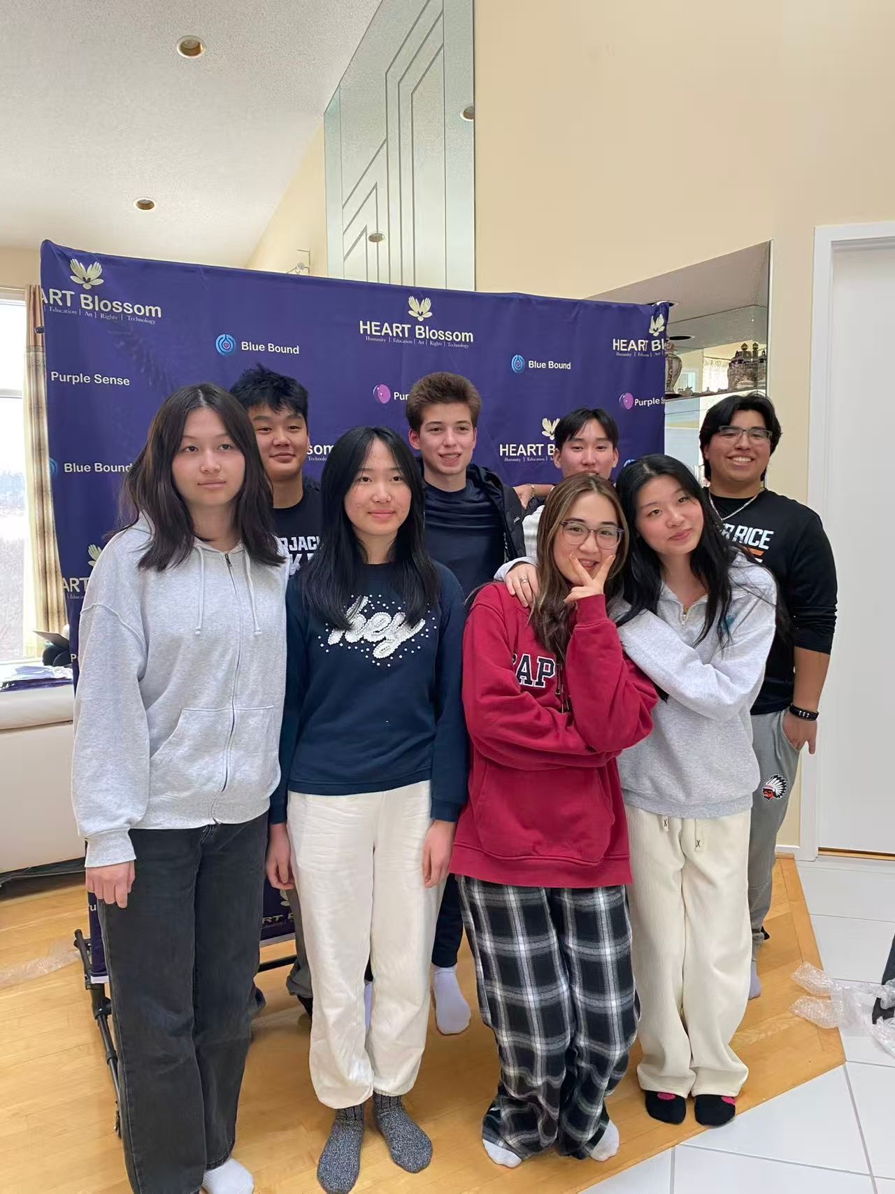 Group of eight diverse teens posing in front of a backdrop with logos, smiling.