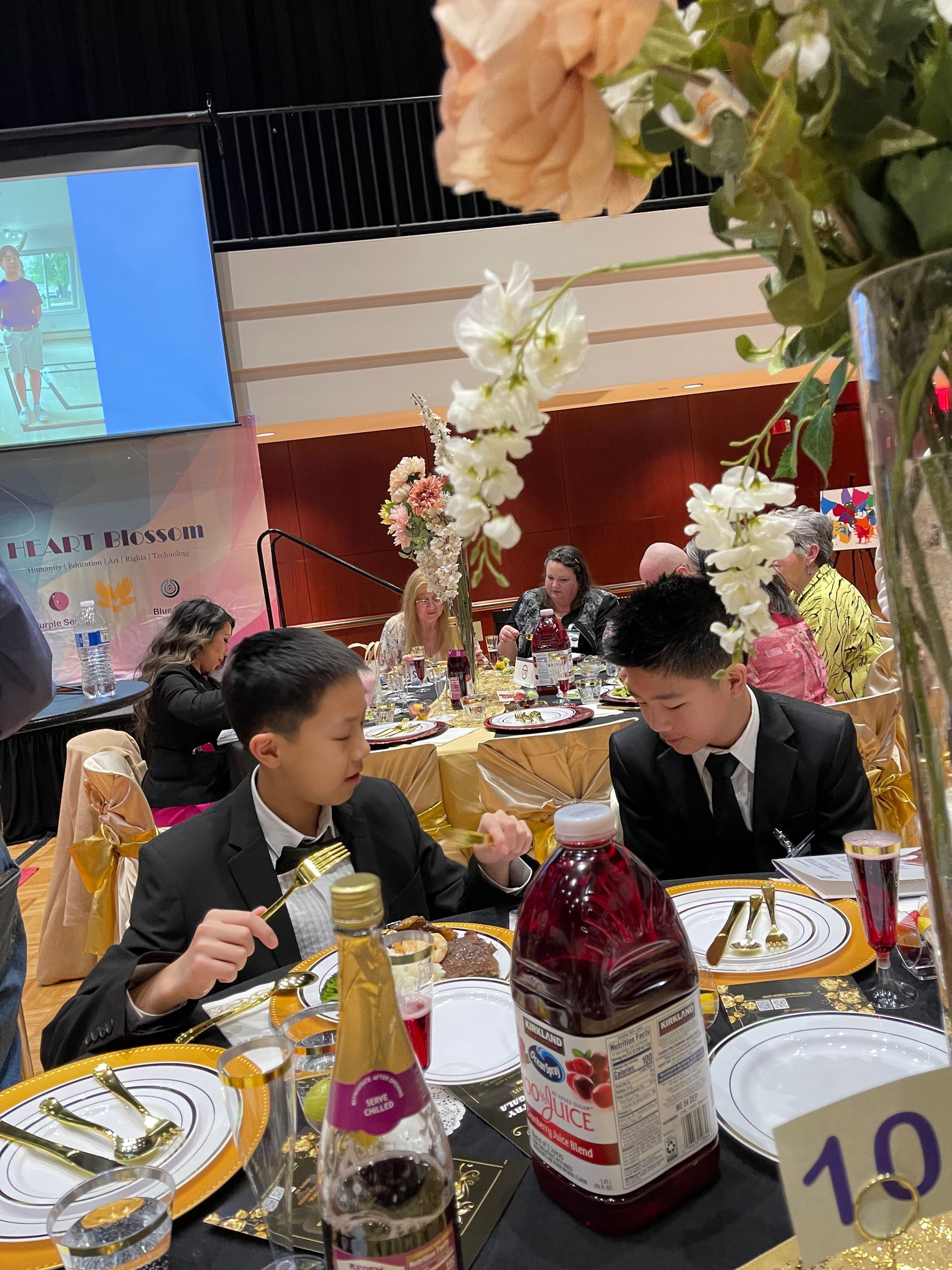 Two young boys in formal attire eating at a banquet table with decorations and guests.
