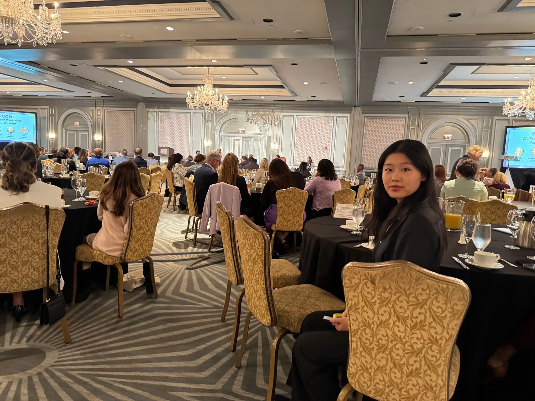 Woman at a conference, looking at camera. People seated at round tables in a large ballroom.