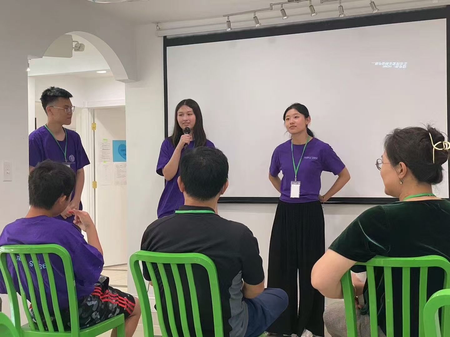 Three young presenters in purple shirts give a presentation; an audience sits in green chairs.
