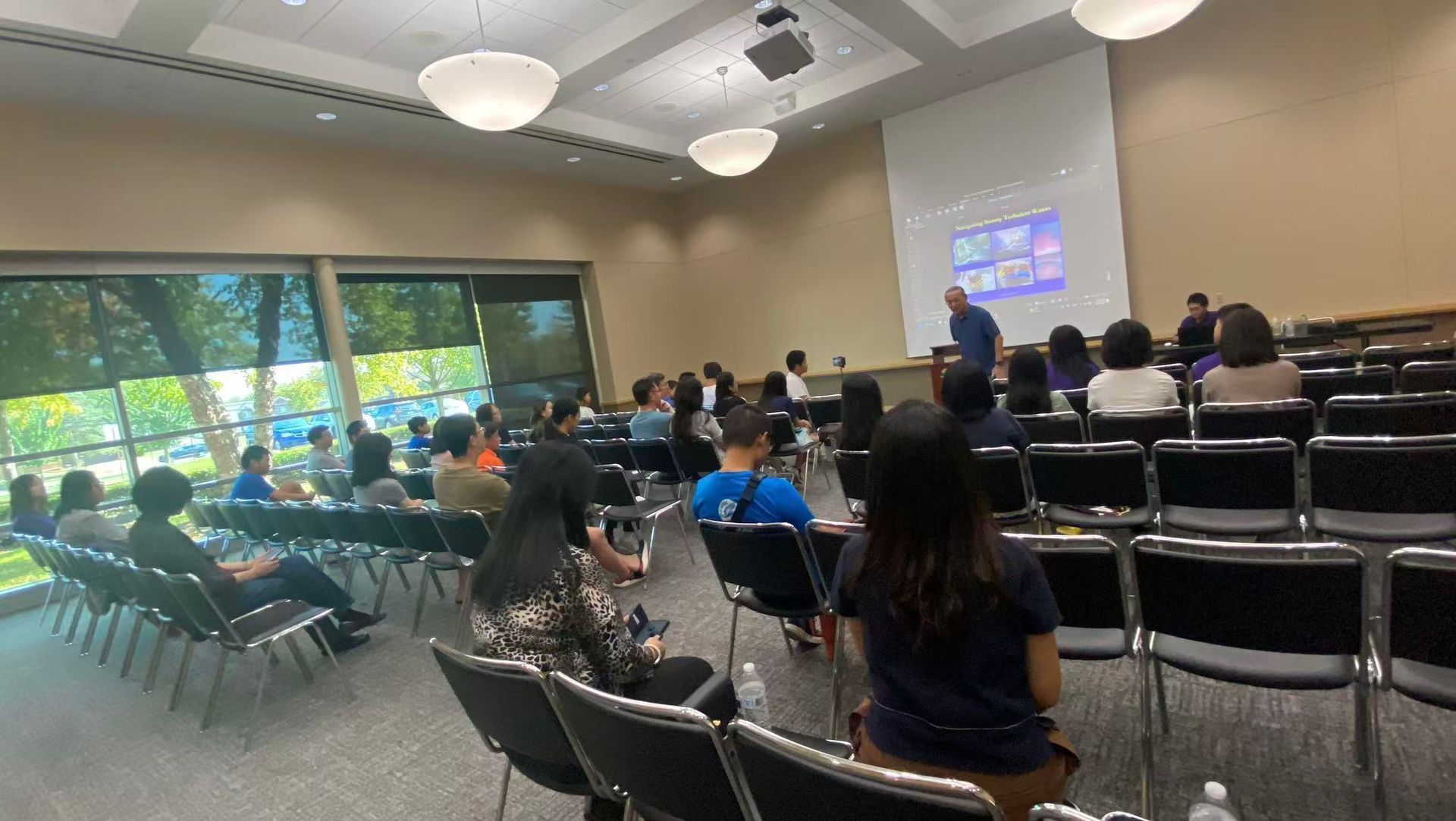 Audience watches a presentation in a lecture hall. A man speaks near a screen. Sunlight streams through a large window.