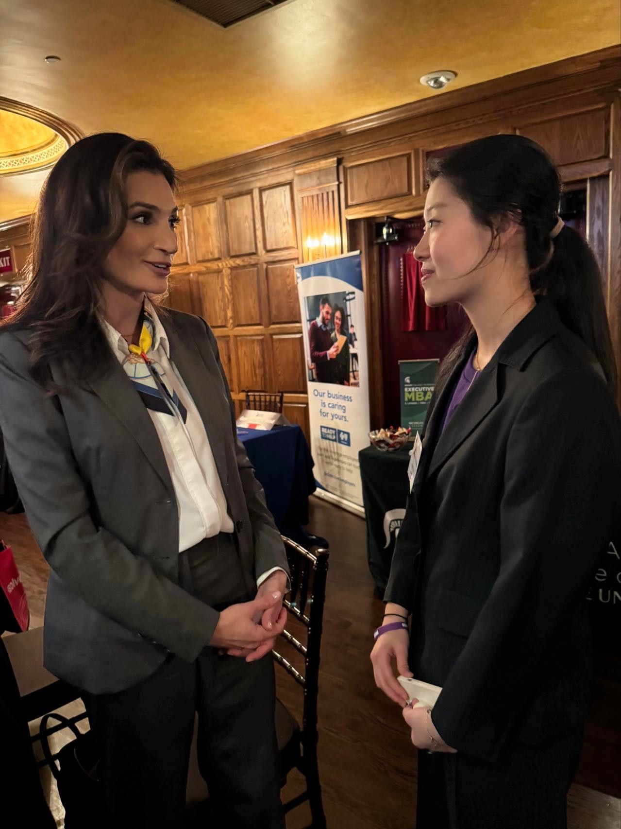 Two women in business suits talking indoors; a banner and chairs are visible in the background.