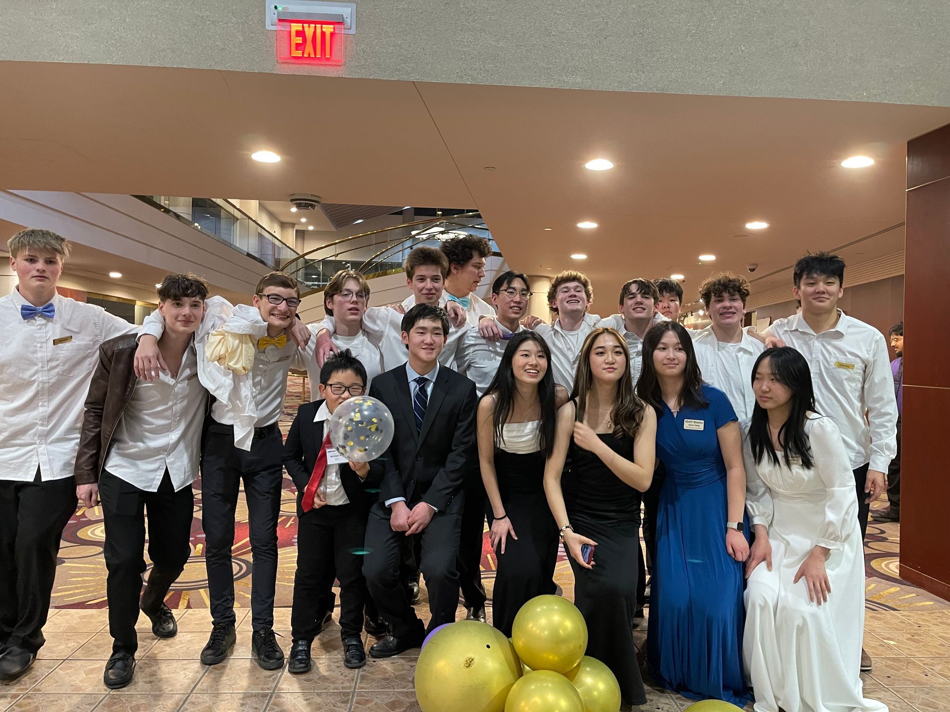 Group of teens in formal attire posing for a photo at an event, smiling and holding gold balloons.