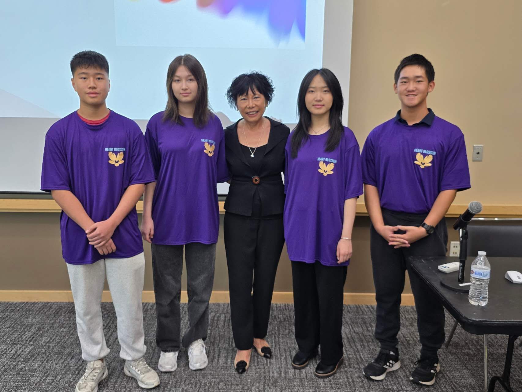 A group of five people smiling. Four teens in purple shirts stand with an older woman in a black suit.