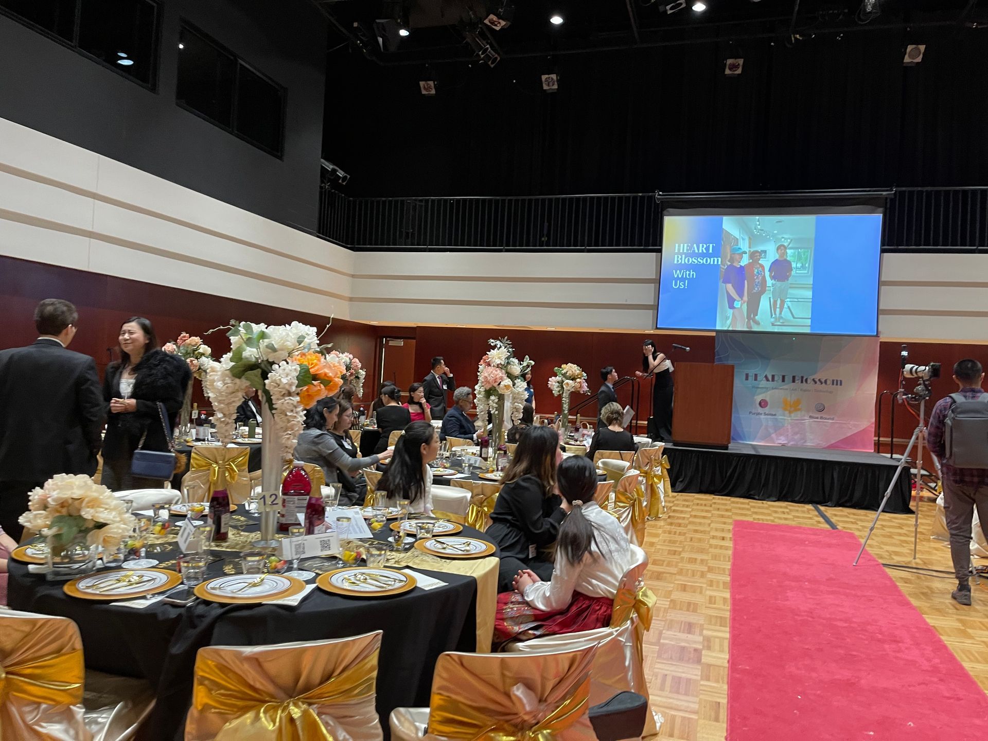 Event in a banquet hall, tables set, people socializing, screen displaying a slideshow.