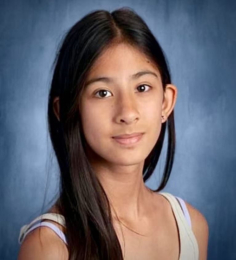 A young woman with long, dark hair and a neutral expression, wearing a tank top, against a blue background.