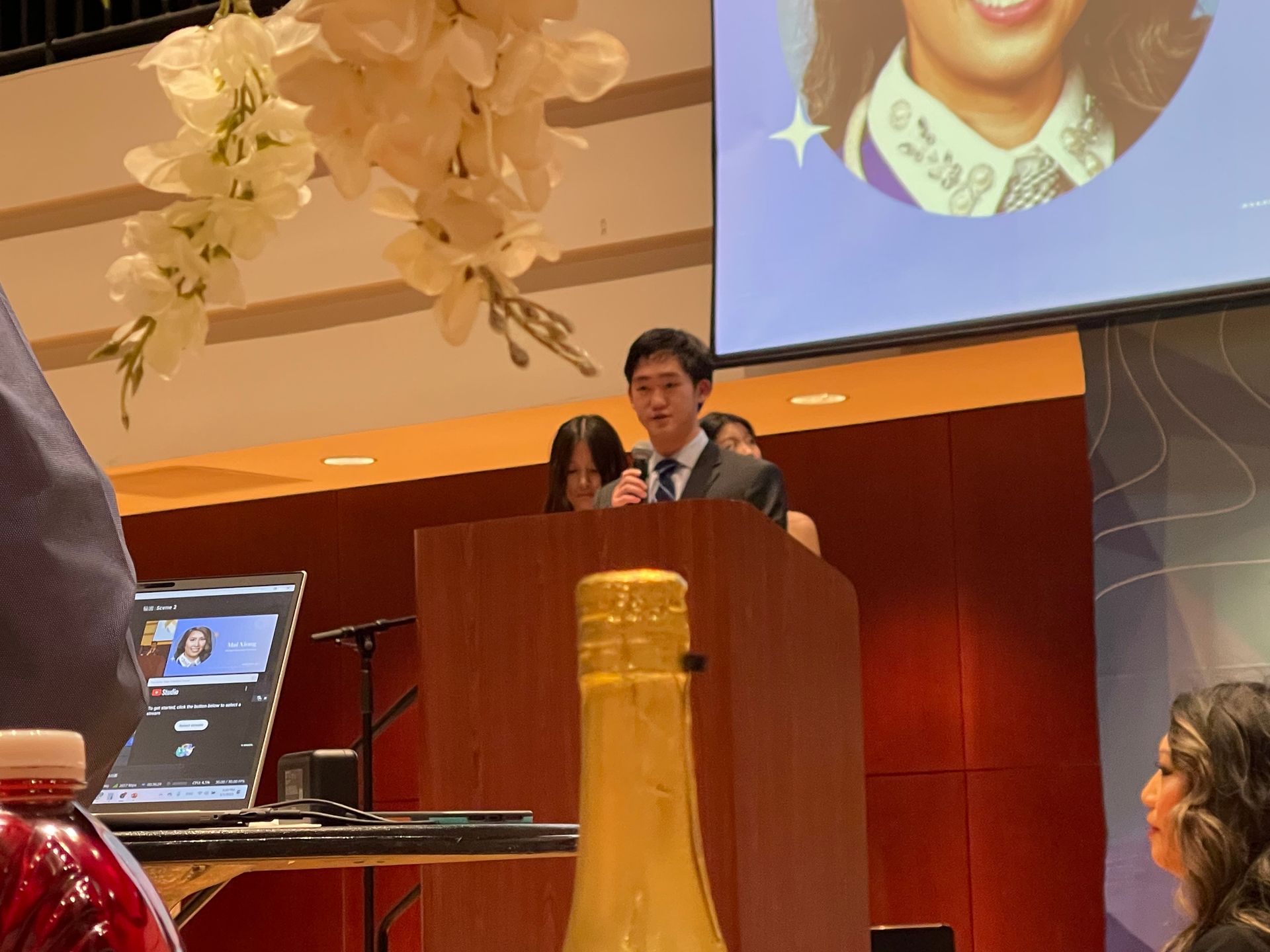 A man speaks at a podium with others behind him, at an event. A screen shows a woman.
