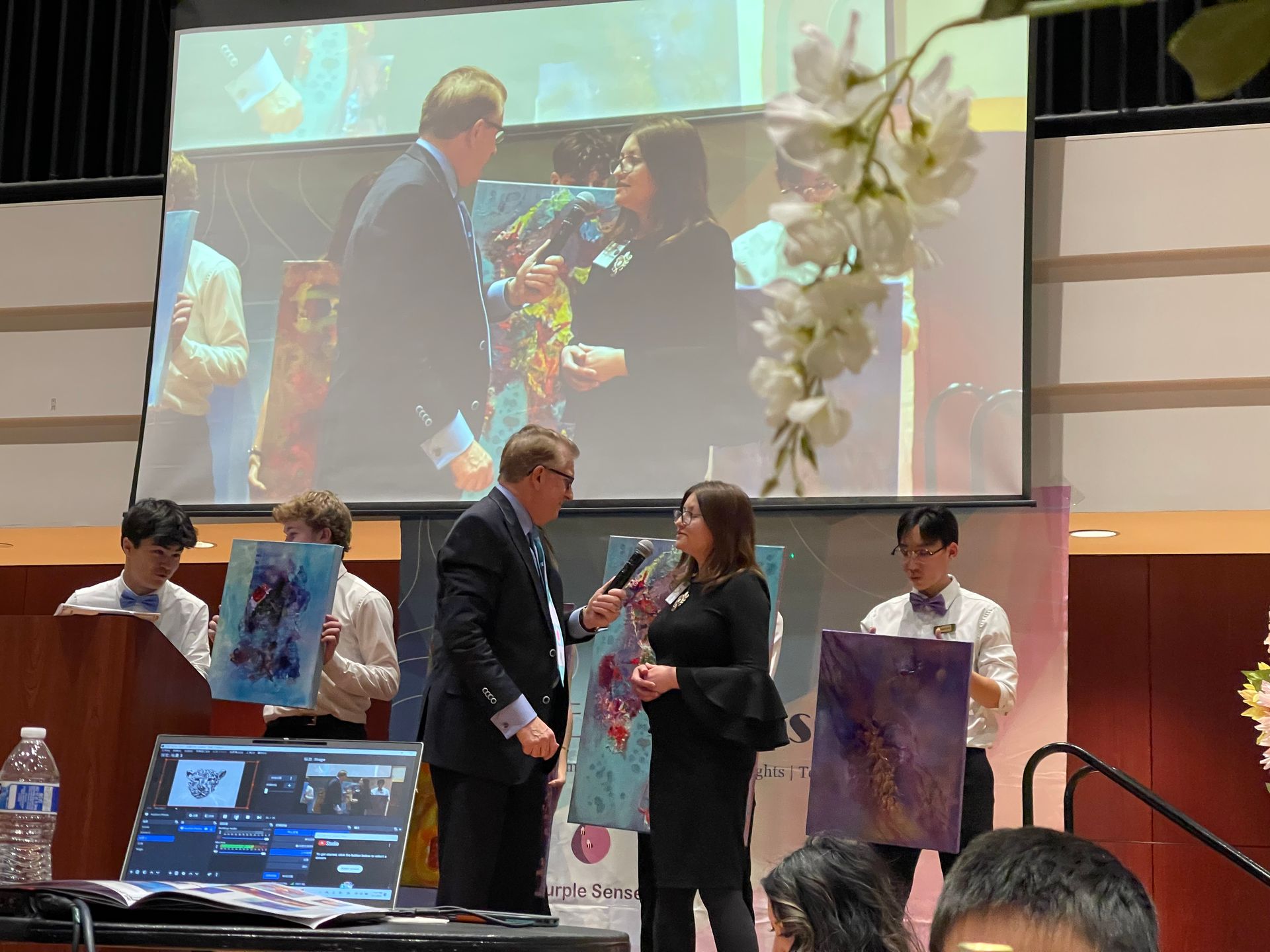 Two people on stage, being interviewed with paintings in background.
