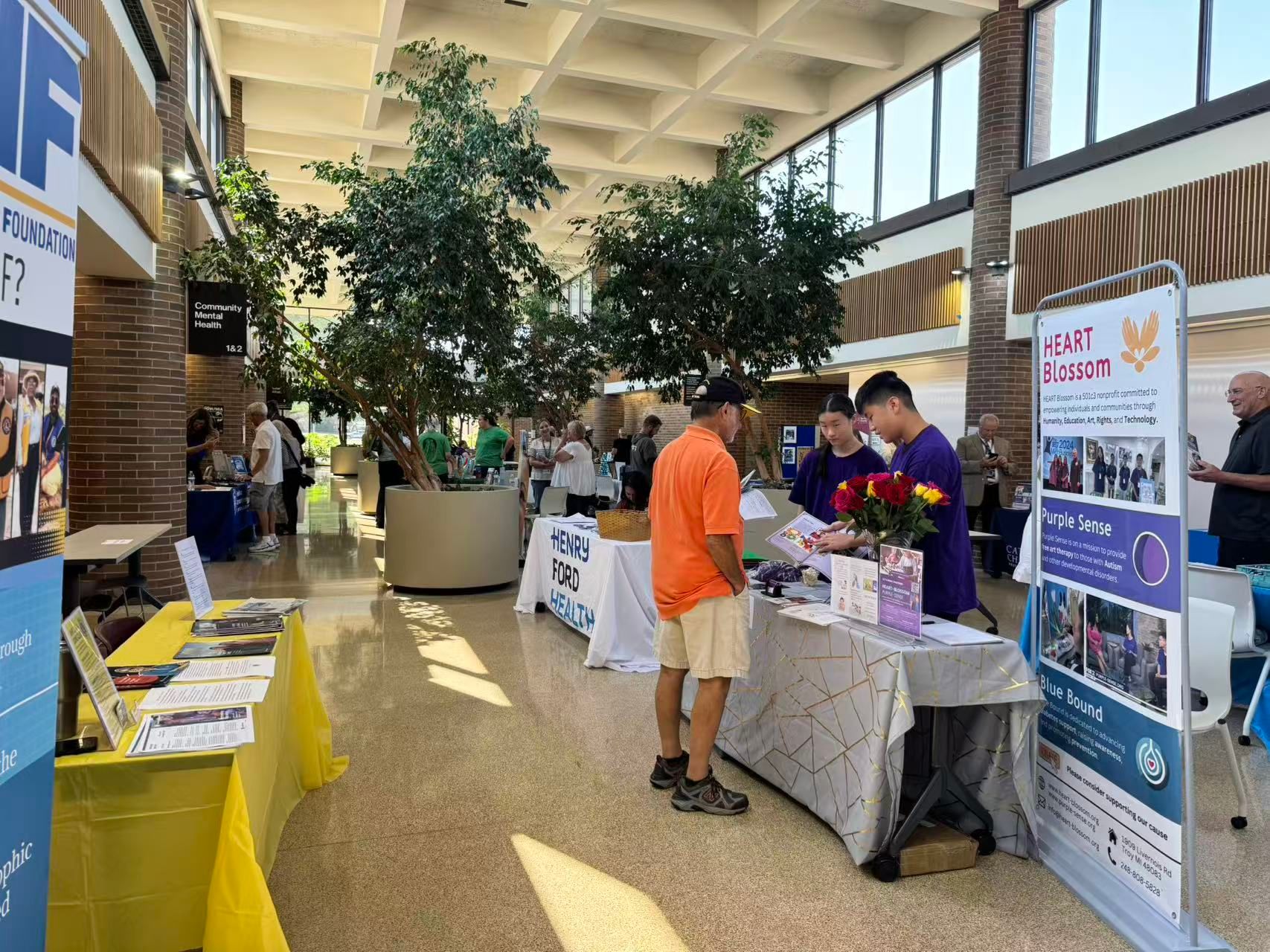 People at information tables inside a building with tall windows and trees in a large pot.