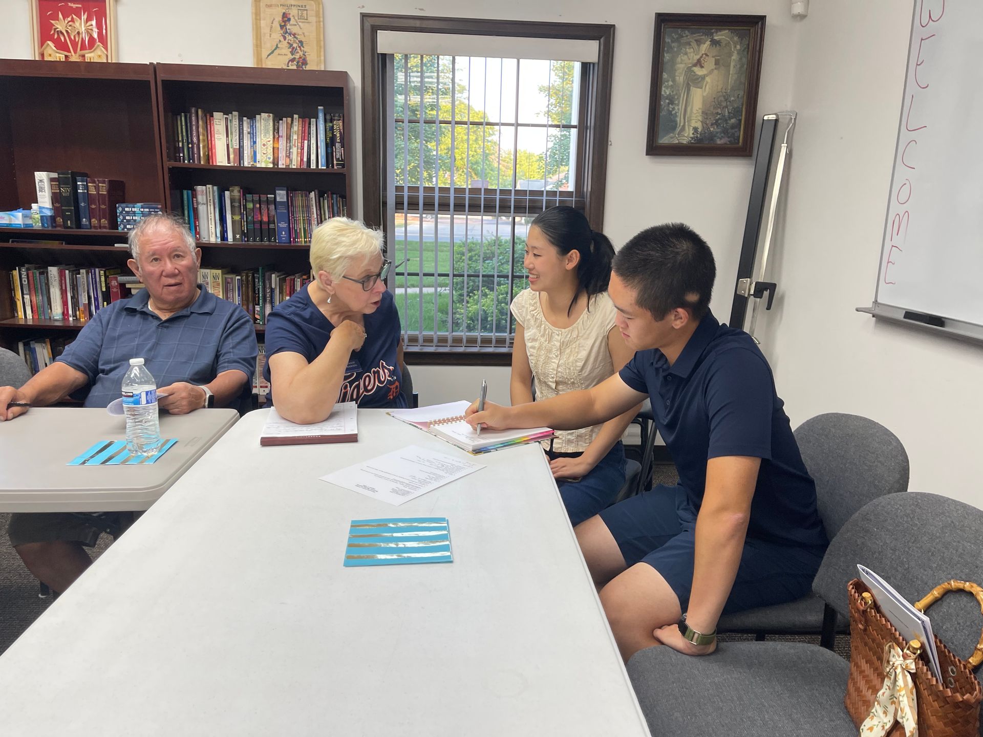Four people at a table, one writing, discussing papers, bookshelves in the background.