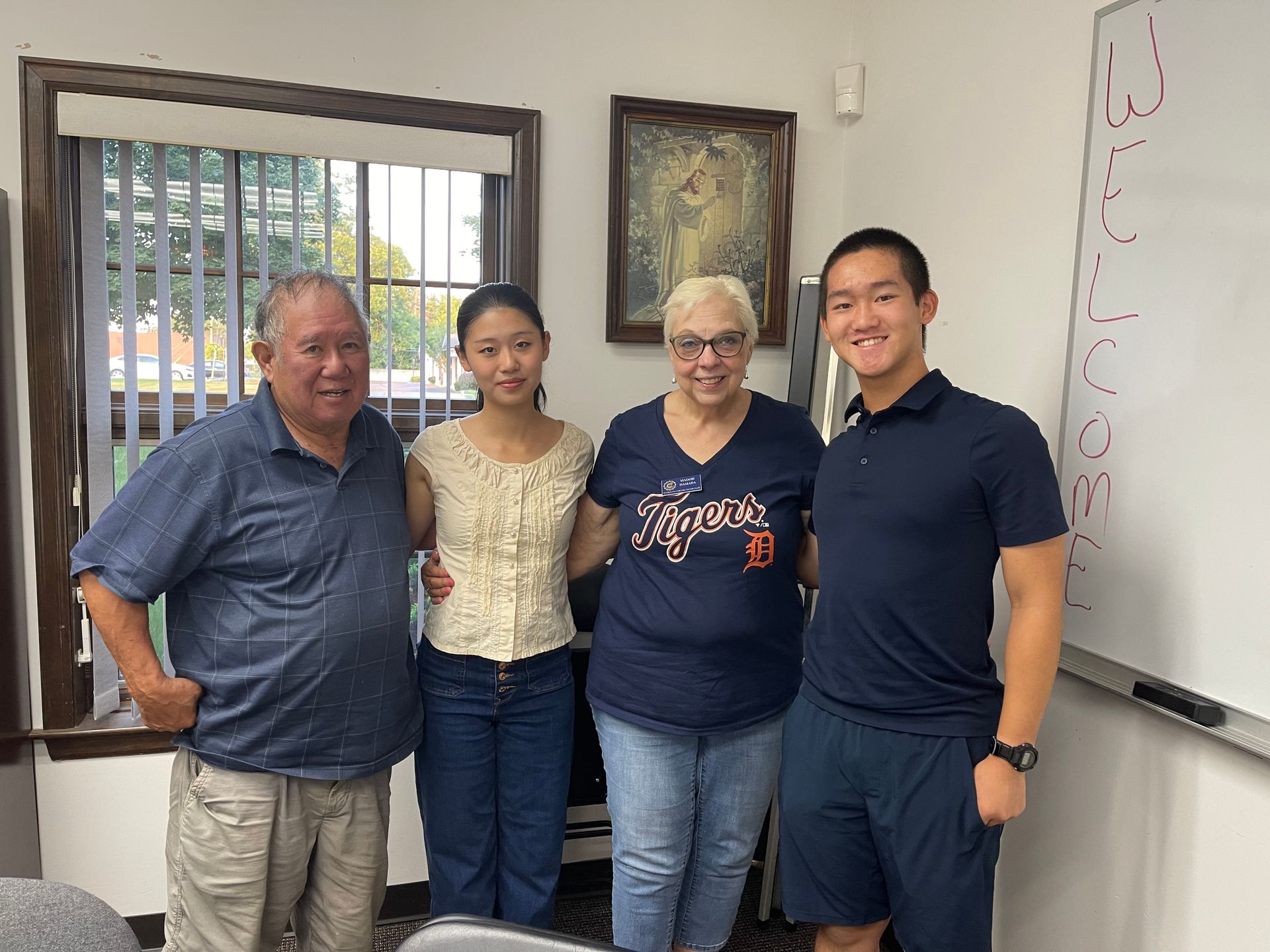 Four people smiling in a room. A window, painting, and whiteboard are visible.