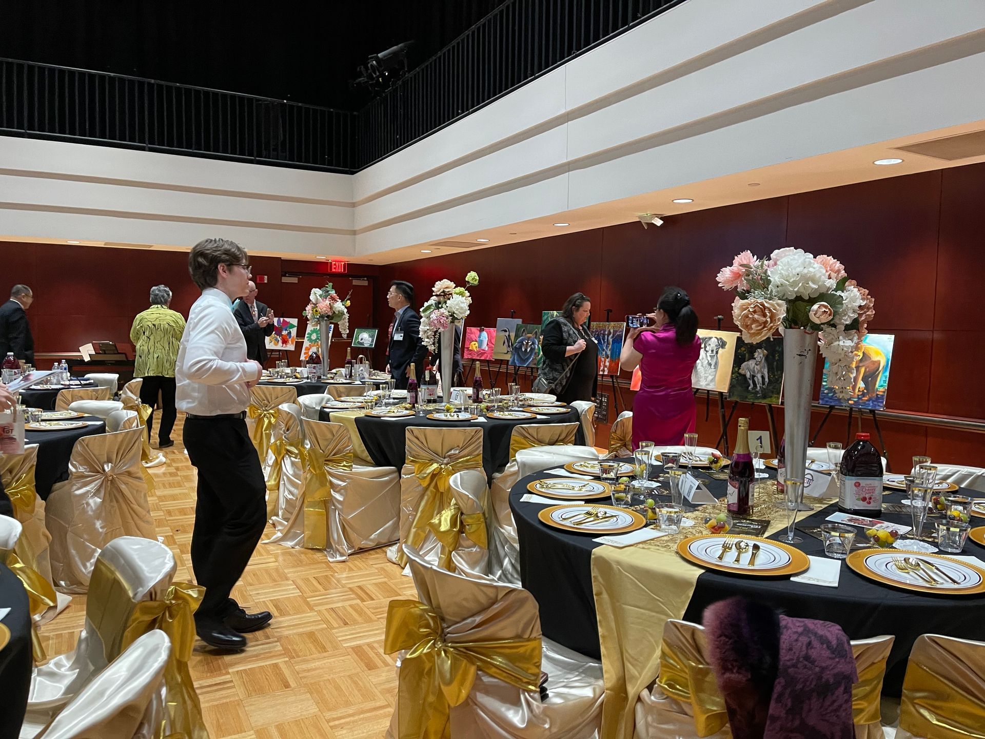 A banquet hall with round tables set for dining; guests browse paintings hung along the wall.