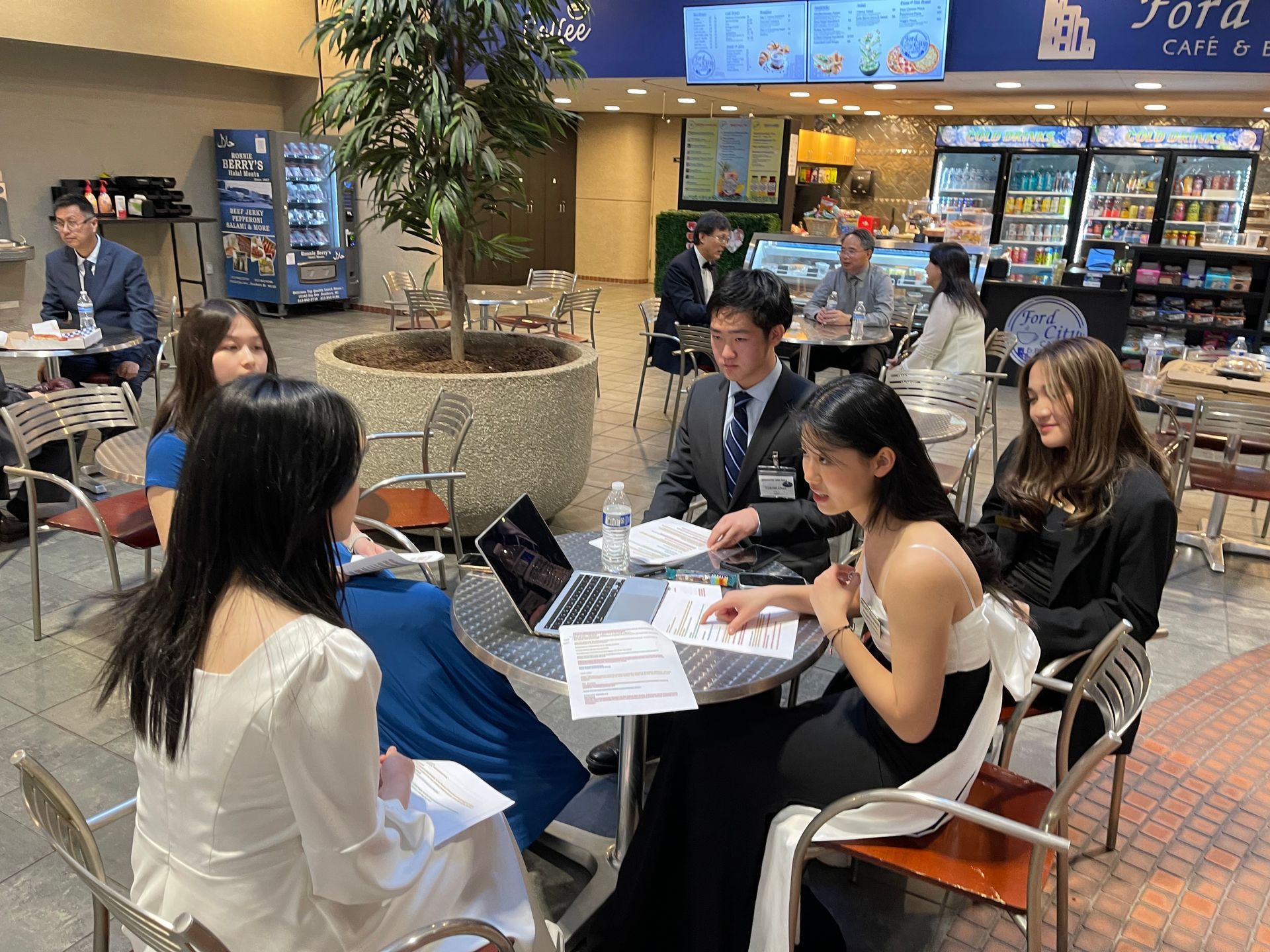 Group of nicely dressed young adults at a table, in a discussion, with laptops and documents in a cafe-like setting.