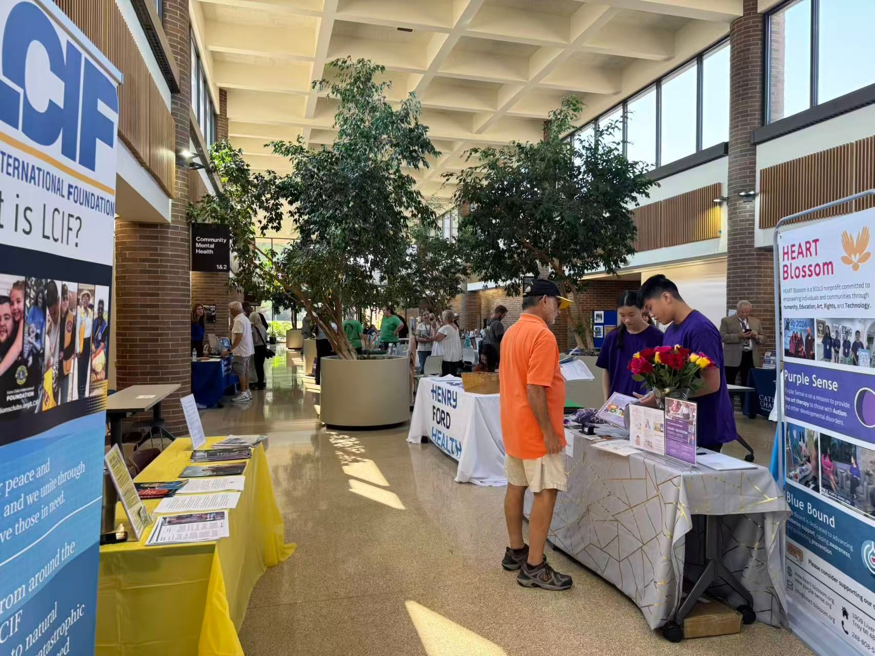 People at tables in a hall with trees, flyers. Person in orange shirt looks at a display. Brick walls, tall ceiling.